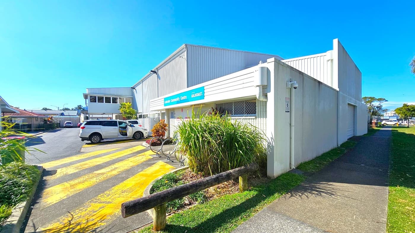 A white building with a blue sign reading "Labrador Community Centre" is on the right. In front, there are green plants and a bicycle rack. A white vehicle is parked on a yellow-striped area to the left. The background shows more buildings and a few parked cars. The ground is a mix of concrete and grass, and a wooden barrier separates the path from the grass. The sky is clear and blue.
