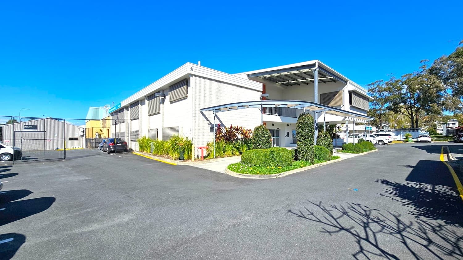 A modern building with a light grey facade and large windows is on the right. It features a covered entrance with neatly trimmed hedges and plants. To the left, a gated area with parked vehicles and an industrial-style building is visible. The foreground shows a wide asphalt driveway with shadows from nearby trees. In the background, more trees and parked cars are visible, adding to the suburban setting.