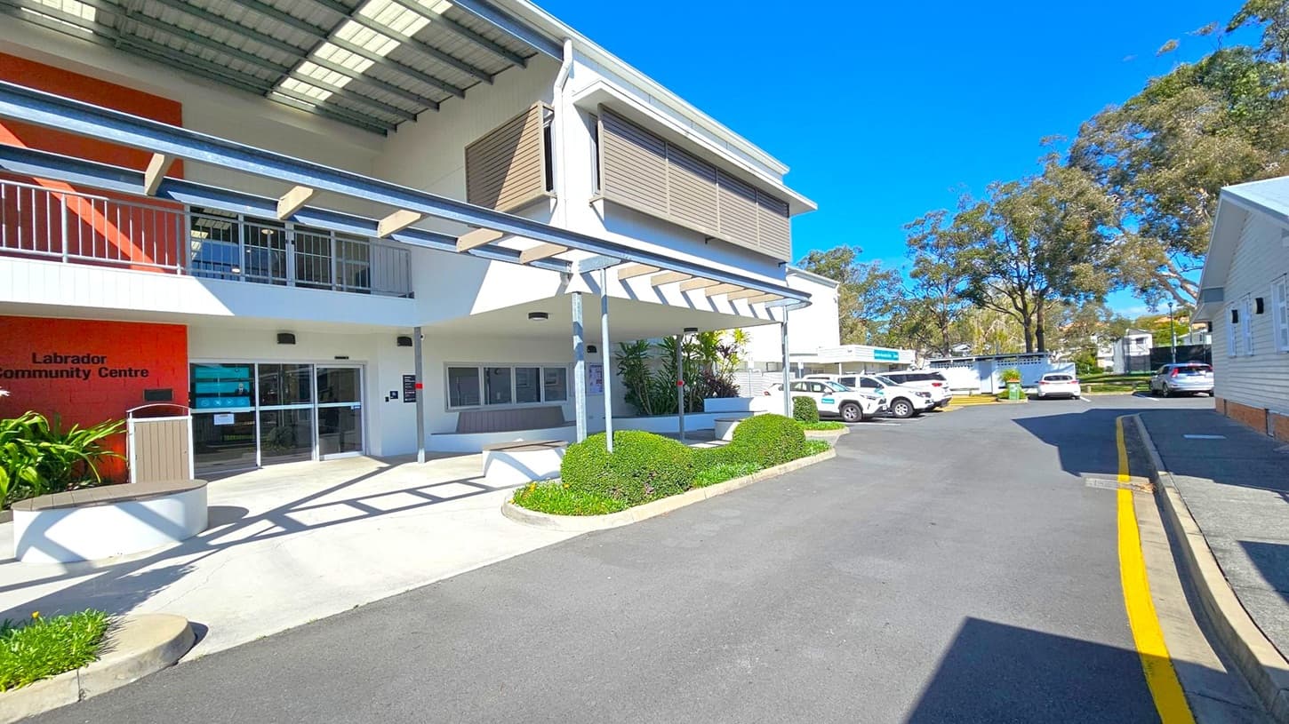 A two-storey building with a modern design, featuring a covered entrance and a sign reading "Labrador Community Centre" on the left. The building has large windows and a metal awning. In the foreground, there are neatly trimmed bushes and a paved walkway. On the right, several parked cars are visible along a driveway, with trees and additional buildings in the background. The sky is clear and blue.
