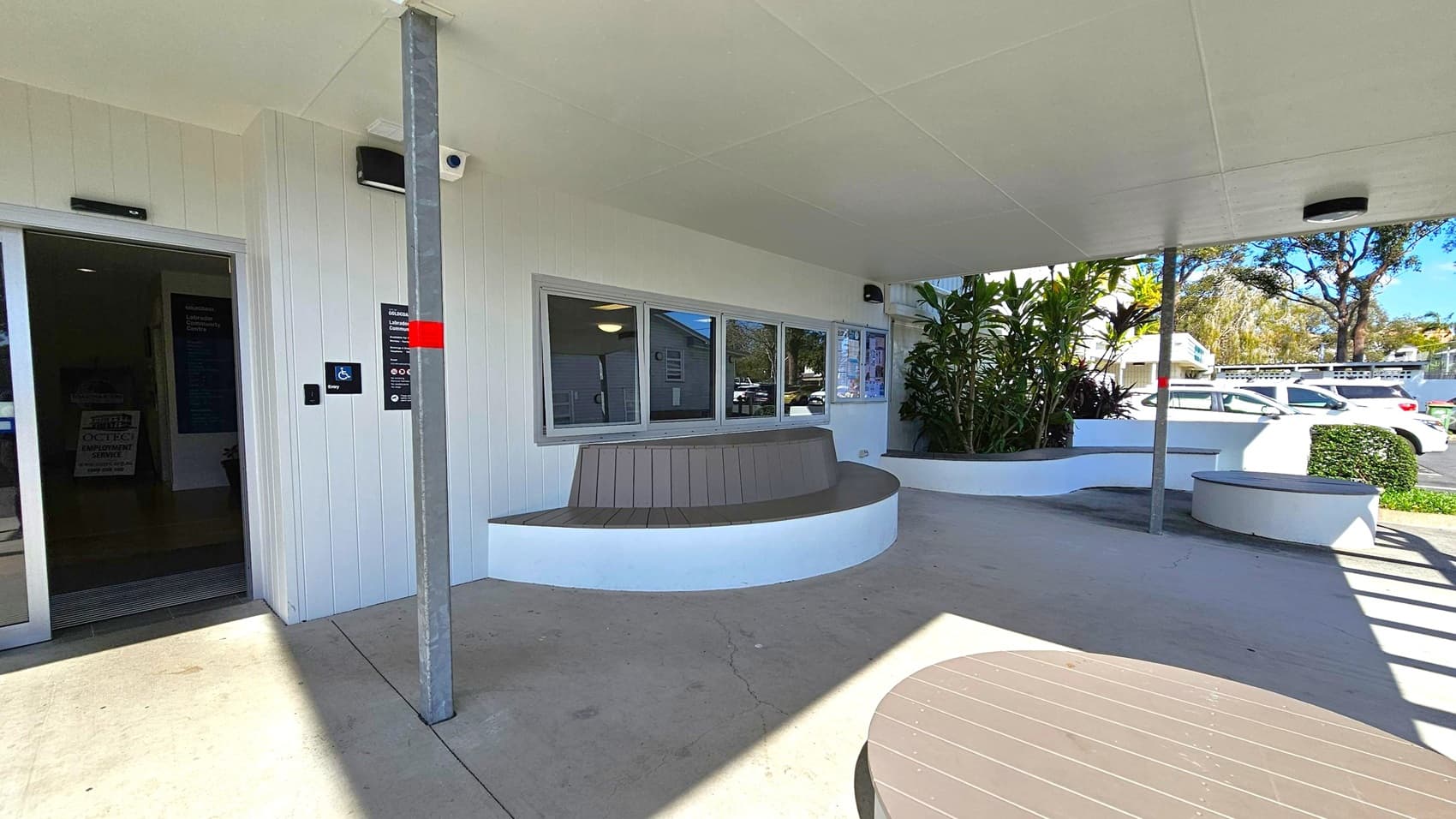 A covered outdoor area with a white ceiling and concrete floor. On the left, there's a dark entrance door with a sign indicating accessibility. In the centre, a curved bench is built into a white wall beneath a window. To the right, a planter with green plants and a small bench is visible. A metal pole supports the ceiling. In the background, several parked cars and trees are visible. A round table is partially visible in the foreground.
