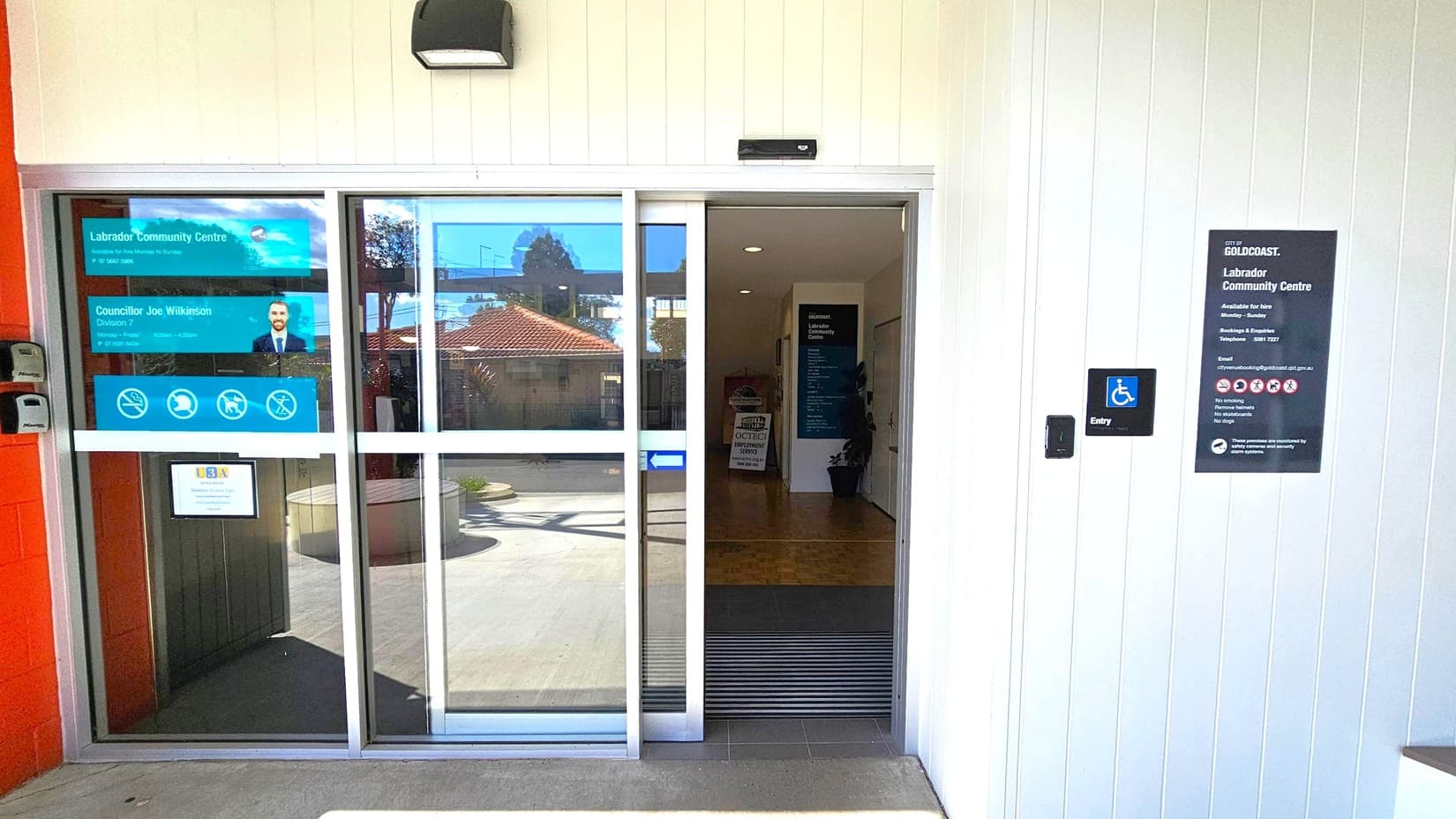 Sliding glass doors at the entrance of a community centre. On the left, signs display the centre's name and contact information, along with a photo of a person. On the right, a wall-mounted sign provides additional details about the centre, including accessibility information. A small blue sign indicates wheelchair access. Inside, a dark hallway is visible with a plant and a sign. The exterior features a light-coloured wall and a concrete floor.