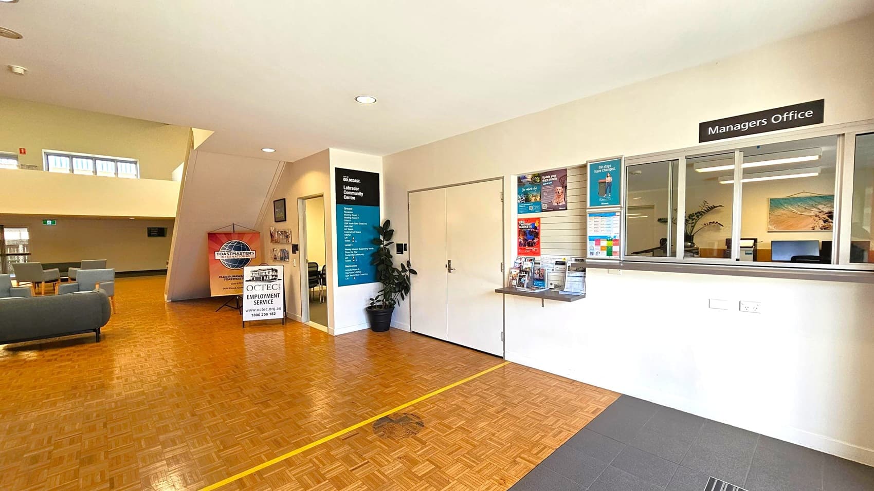 A spacious interior with a parquet floor. On the right, there's a "Managers Office" with a window and various posters. In the centre, a door is flanked by a plant and a sign listing services. On the left, a seating area with grey chairs and a table. A sign for "Toastmasters" and "CCTEC Employment Service" is visible. The ceiling is light-coloured with recessed lighting, and a staircase leads to an upper level with windows.