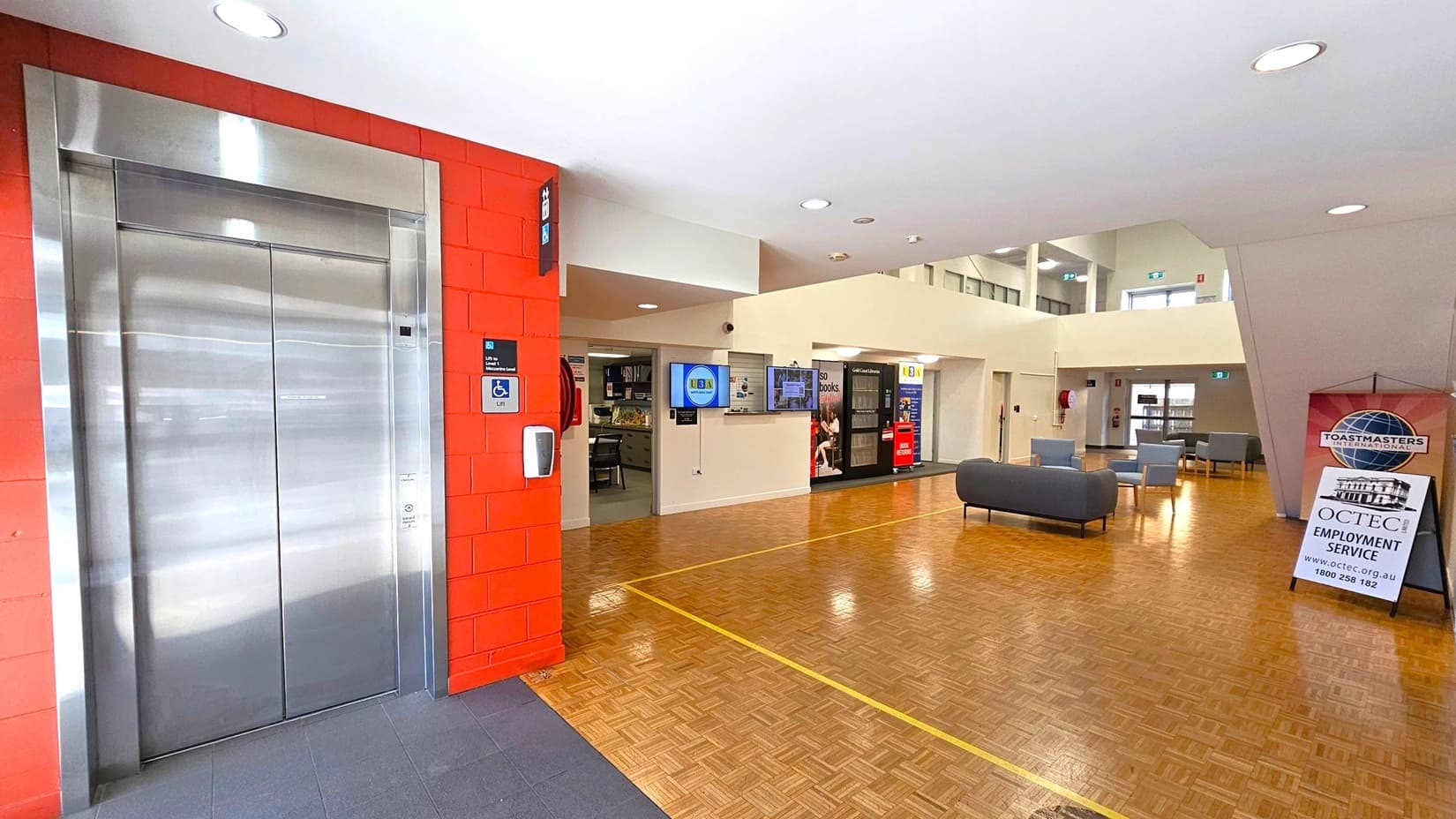 On the left, there is a stainless steel lift with a red brick wall. A sign indicates accessibility features. In the centre, a digital screen is mounted on a cream wall, with a small office area visible. The floor is wooden with a yellow line. On the right, there are grey sofas and chairs, and a sign for OCTEC Employment Service. The ceiling is white with recessed lighting.