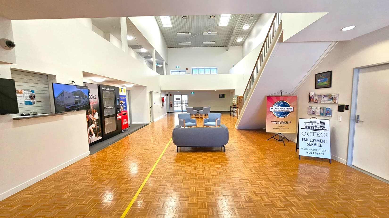 A spacious interior with a parquet floor and high ceiling. On the left, there's a counter with a TV screen and a vending machine. In the centre, a seating area with a sofa and chairs. On the right, a staircase leads to an upper level. Signs for Toastmasters International and OCTEC Employment Service are displayed near the wall, along with a brochure rack. The ceiling features skylights and recessed lighting.
