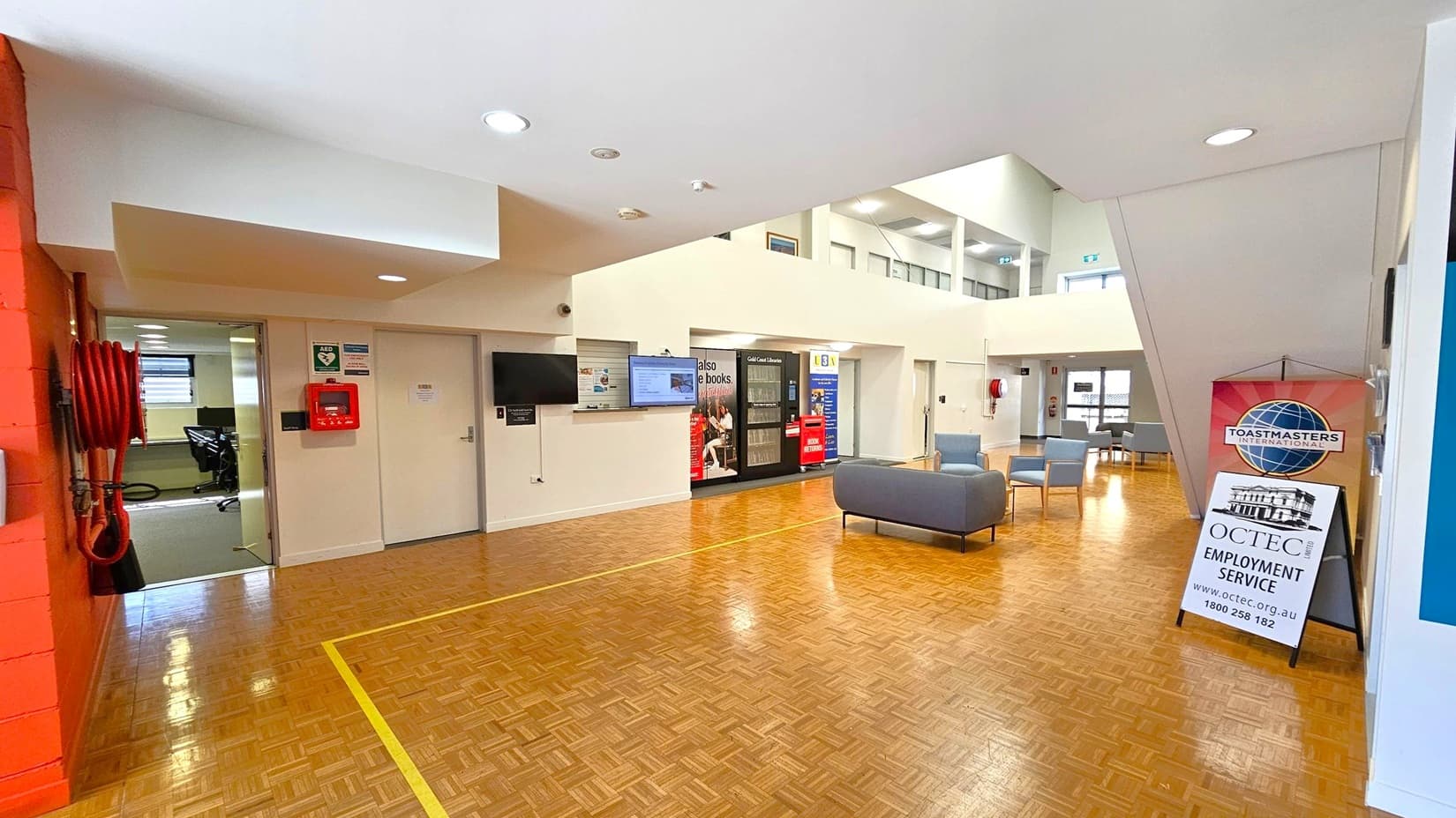 A spacious interior with a parquet wooden floor and a white ceiling with recessed lights. On the left, a red fire hose and a partially open door leading to an office area. In the centre, a wall-mounted TV and a vending machine. On the right, a seating area with grey chairs and a sign for Toastmasters International and OCTEC Employment Service. The background features large windows and additional seating.