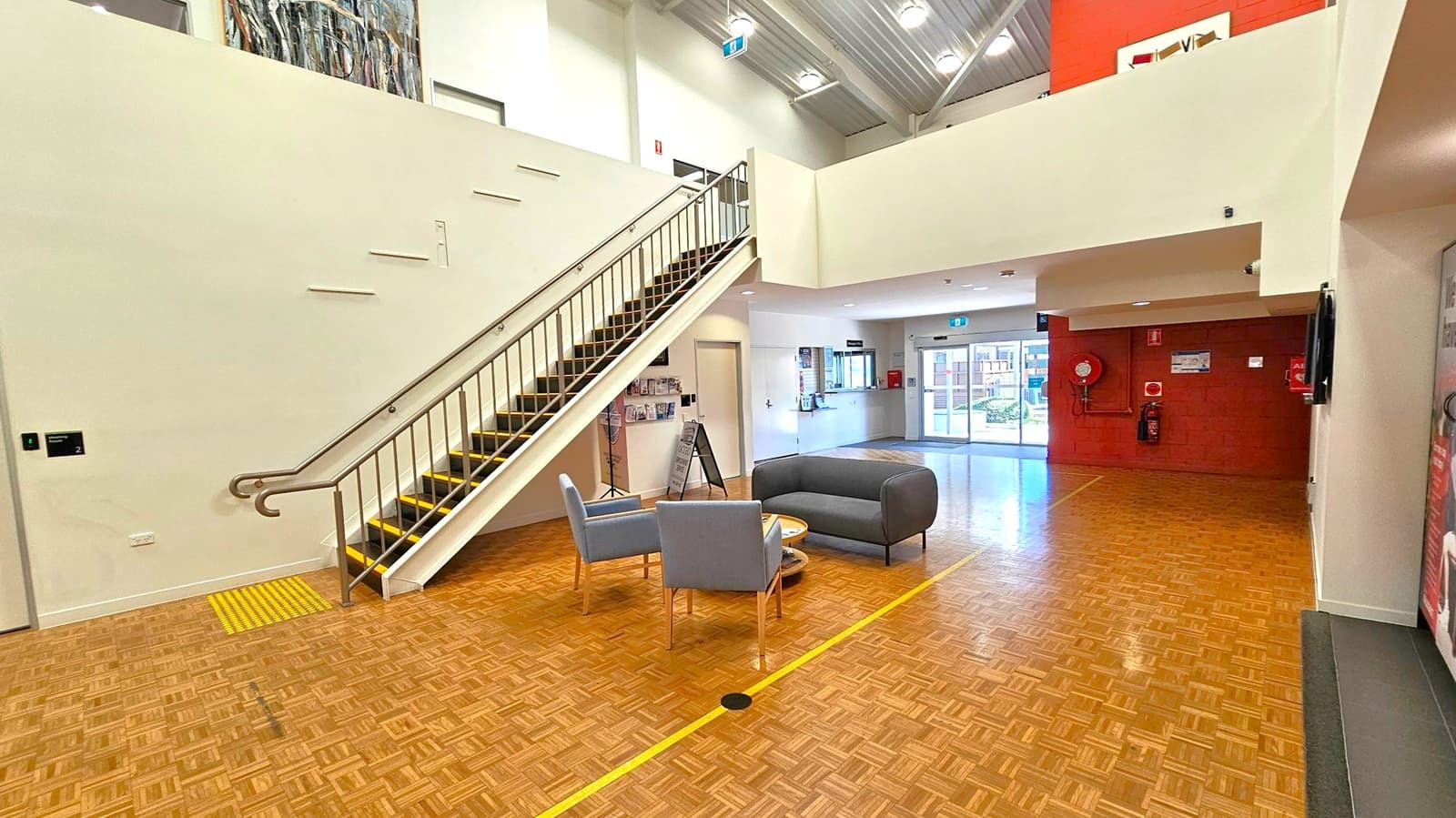 Spacious interior with a parquet floor. On the left, a staircase with a metal railing leads to an upper level. Below the stairs, a wall-mounted display holds brochures. In the centre, a seating area with two grey chairs and a small table. The right side features a red wall with a fire extinguisher and a hose reel. The background shows a reception area with a window and a glass door leading outside. The ceiling is high with exposed beams and lights.