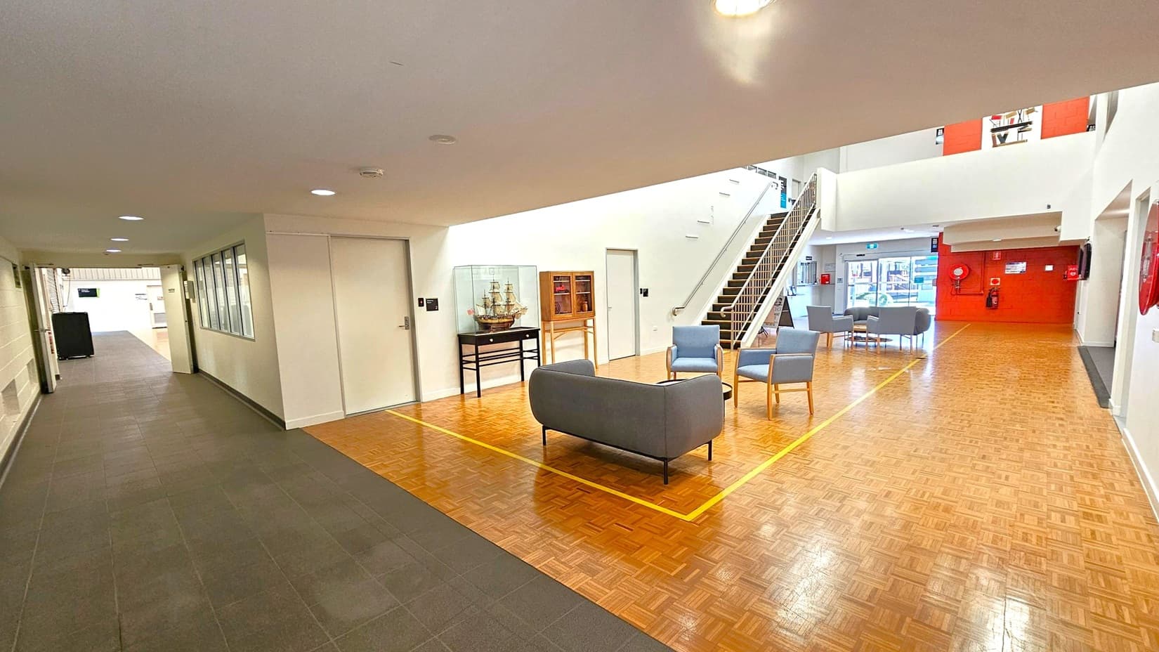 A spacious interior with a parquet floor. On the left, a hallway with dark tiles and a row of windows. In the centre, a seating area with a grey sofa and blue chairs around a small table. A model ship in a glass case and a wooden cabinet are against the wall. A staircase with a metal railing leads to an upper level. On the right, more seating and a red wall with fire safety equipment. The ceiling is white with recessed lighting.