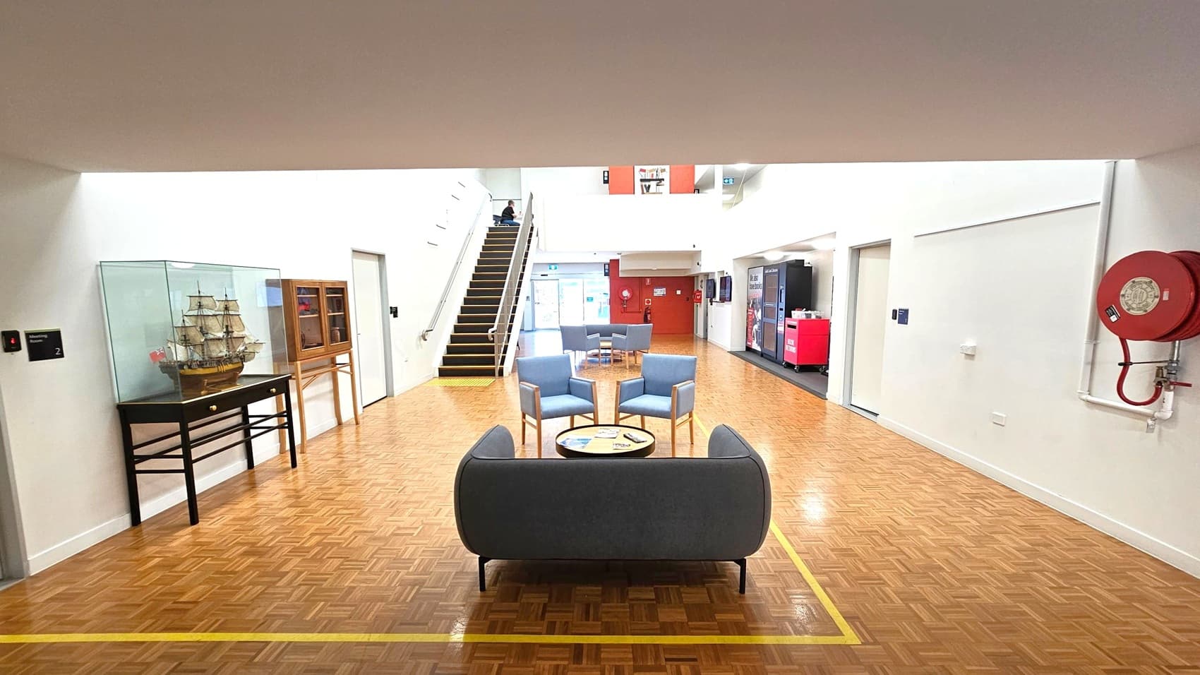 A spacious indoor area with a parquet floor. In the foreground, a seating arrangement includes a grey sofa and three blue chairs around a circular table. On the left, a model ship is displayed in a glass case on a black table. A staircase leads up to the left, and a fire hose reel is mounted on the right wall. In the background, vending machines and a red wall are visible, with more seating near a glass entrance. The ceiling is plain and white.