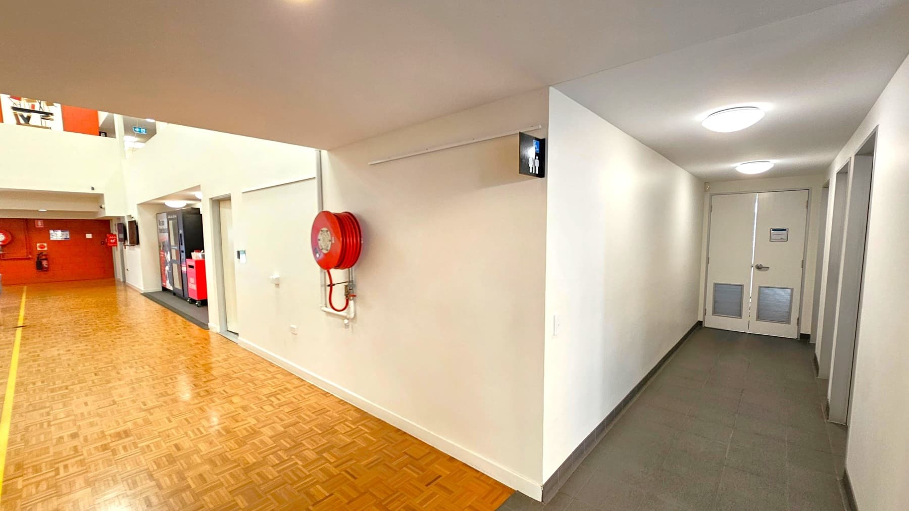 A hallway with a wooden parquet floor on the left and a tiled floor on the right. A red fire hose reel is mounted on the wall near the corner. A sign with male and female figures indicates a restroom down the corridor on the right. The ceiling is white with circular lights. In the background, there are vending machines and a red wall with fire safety equipment.