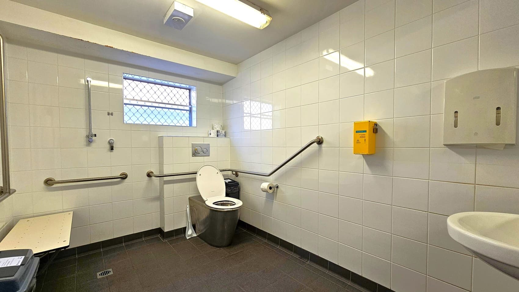 A public restroom with white tiled walls and a dark tiled floor. In the centre, a toilet with a raised seat and a lid is flanked by metal grab bars. Above the toilet, a small window with a wire mesh cover allows light in. To the right, a yellow sharps disposal box and a soap dispenser are mounted on the wall. A sink is partially visible in the foreground on the right. On the left, a foldable bench and a waste bin are present. The ceiling has a fluorescent light fixture.