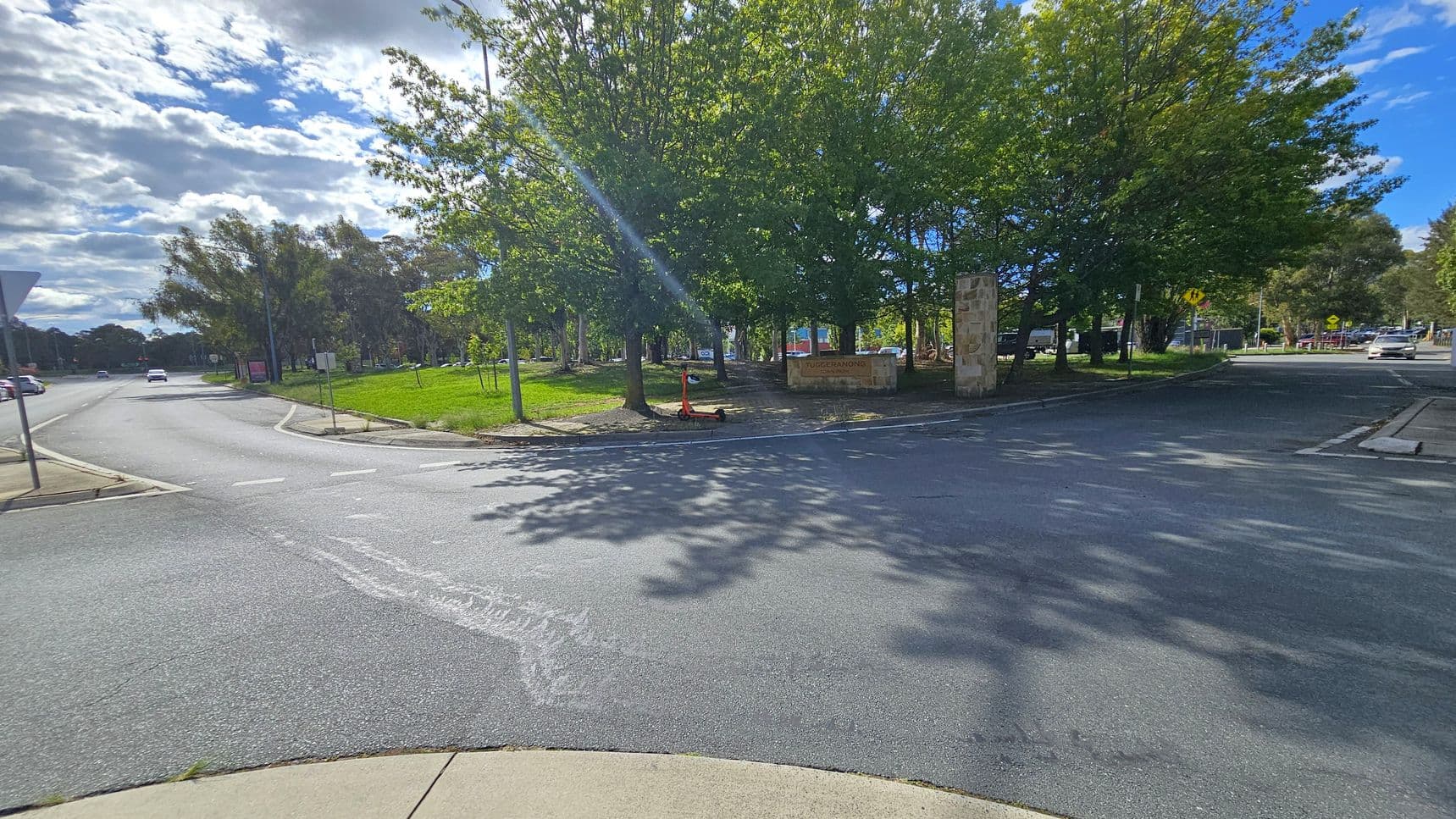 A street intersection with a road curving to the left and another road extending to the right. In the centre, there are several large trees providing shade, with a sign partially visible among them. A scooter is parked near the trees. The pavement is smooth, and the area is well-lit with sunlight filtering through the leaves. On the left, a grassy area is bordered by a footpath, and a few cars are visible in the distance.