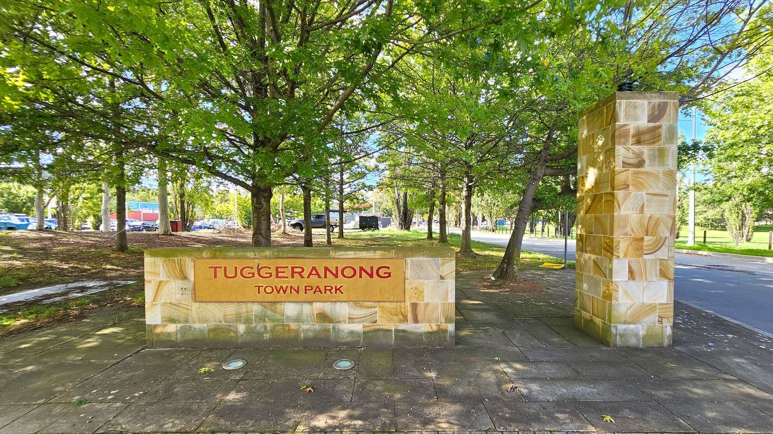 A stone sign with "Tuggeranong Town Park" in red letters is on the left, set against a backdrop of lush green trees. To the right, a tall stone pillar stands beside a paved path. The ground is covered with square tiles, and a road runs parallel to the path. In the background, more trees and a few parked cars are visible.