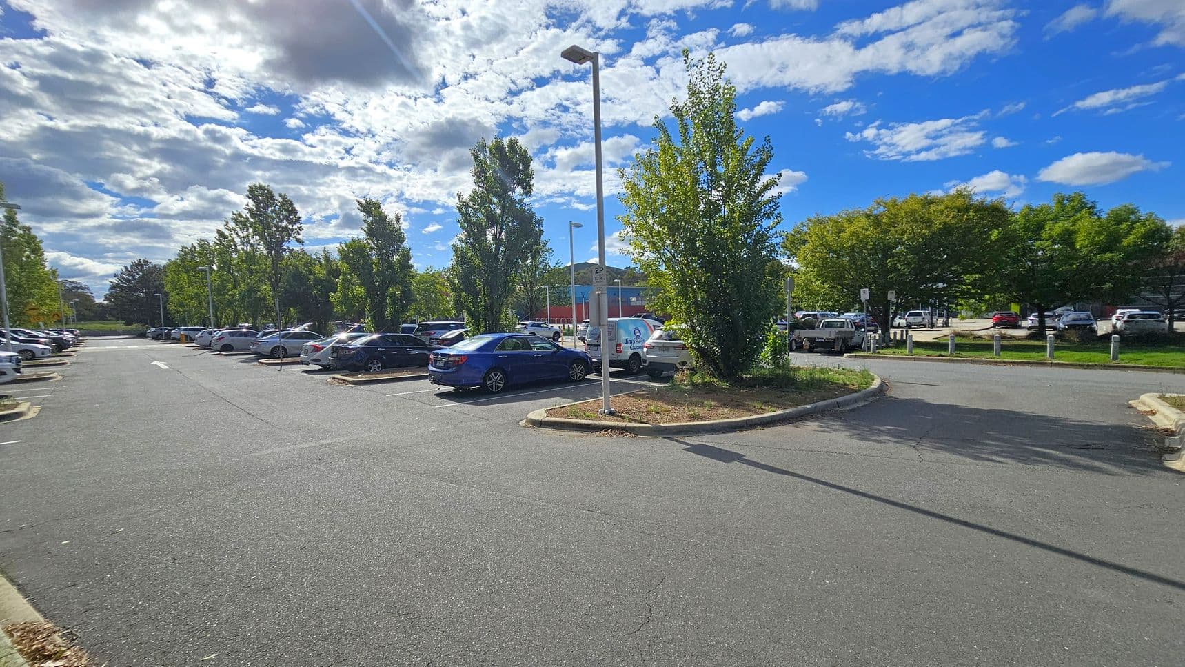 A car park with several vehicles is visible, surrounded by trees. On the left, there are rows of parked cars and tall streetlights. In the centre, a blue car is parked near a small island with a tree and a signpost. On the right, more cars are parked, and a building with a blue and red facade is partially visible in the background. The ground is paved with asphalt, and the area is well-lit by daylight.