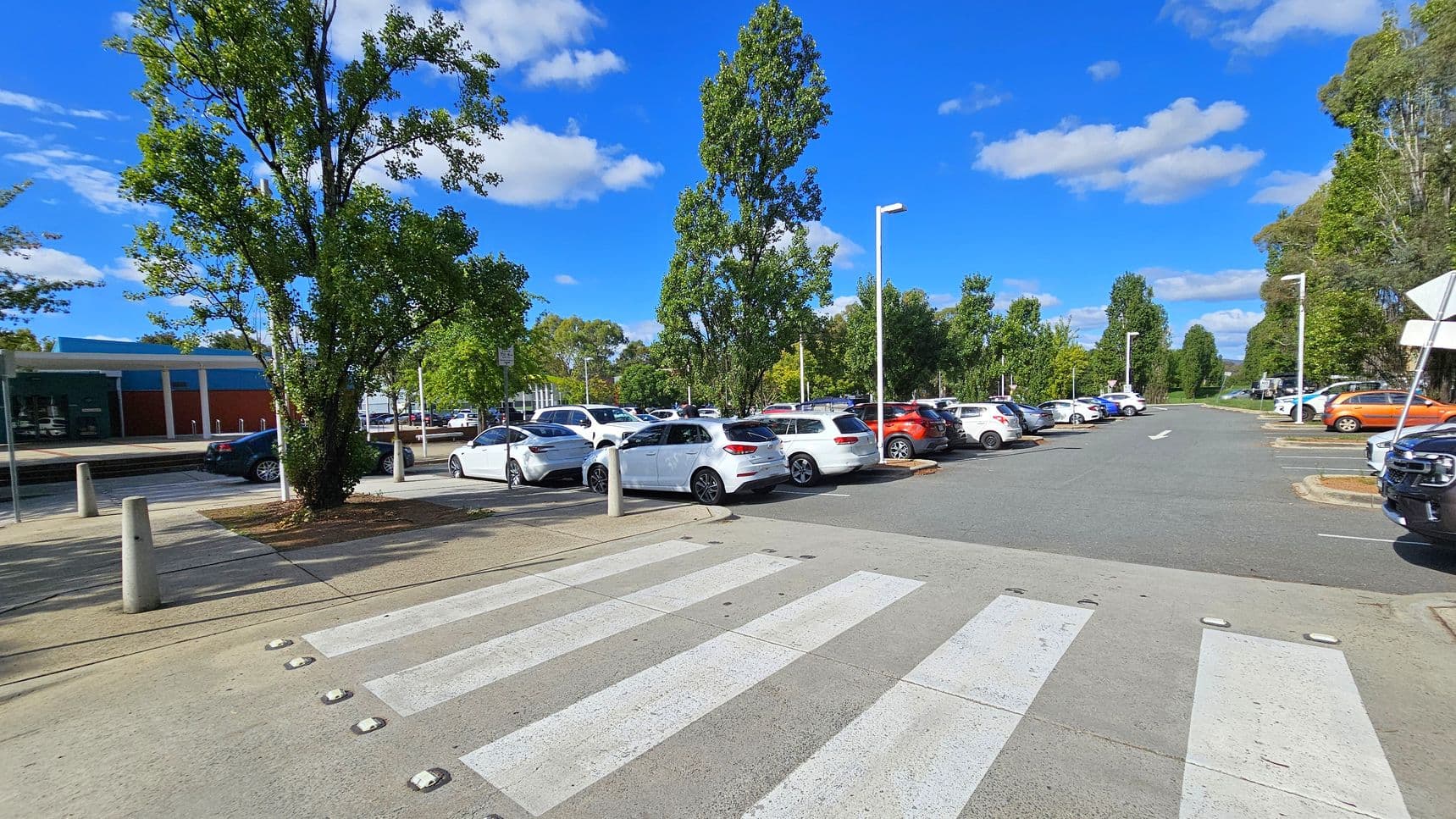 A car park with a zebra crossing in the foreground. On the left, there is a large tree and a building with a blue and red facade. Several cars are parked, mostly white, with a few in other colours like orange. Tall trees line the parking area, and street lamps are visible. The road extends into the distance, flanked by more trees and greenery. The sky is clear with a few clouds.