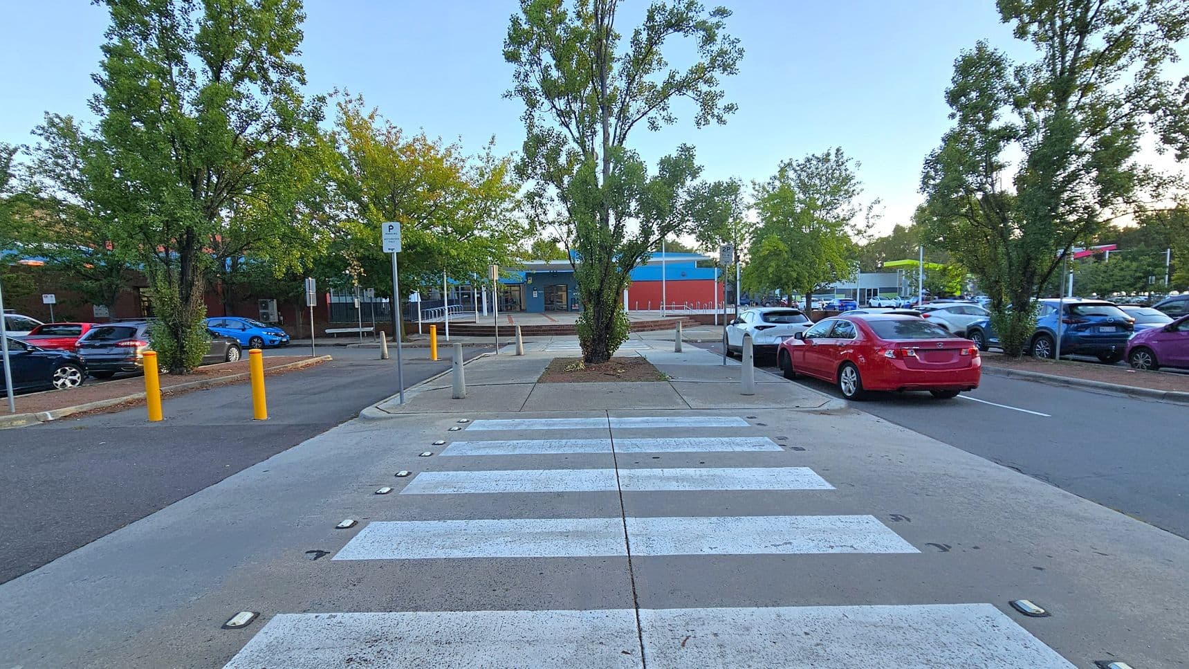 A zebra crossing leads to a tree-lined area with a central tree. On the left, there are parked cars and yellow bollards along the road. On the right, a red car is parked near more trees and additional vehicles. In the background, a building with blue and red accents is visible, surrounded by more trees and a parking area. The ground is paved, and the scene is well-lit.