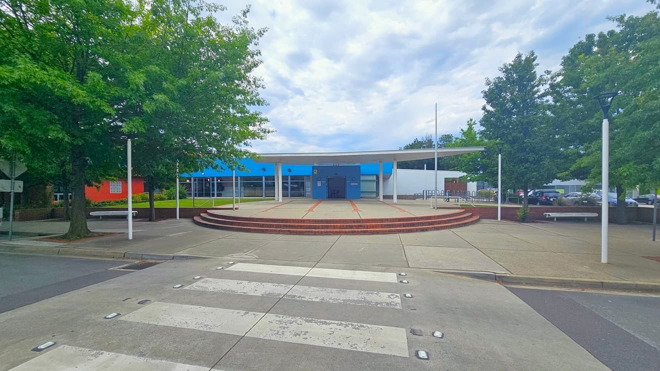 A building with a blue and white facade is in the centre, featuring a circular entrance area with steps leading up to it. The foreground has a zebra crossing on a paved surface. On the left and right, there are large green trees and white lamp posts. The area is open and spacious, with a few benches visible near the trees.
