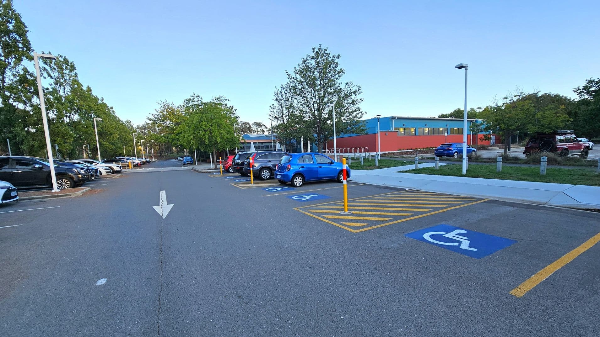 A car park with a row of parked cars on the left, including black and silver vehicles. On the right, there are designated disabled parking spaces marked with blue signs and yellow lines. A blue car is parked in one of these spaces. In the background, a building with blue and orange walls is visible, surrounded by trees. The ground is paved, and there are streetlights along the parking area. An arrow is painted on the road, indicating direction.