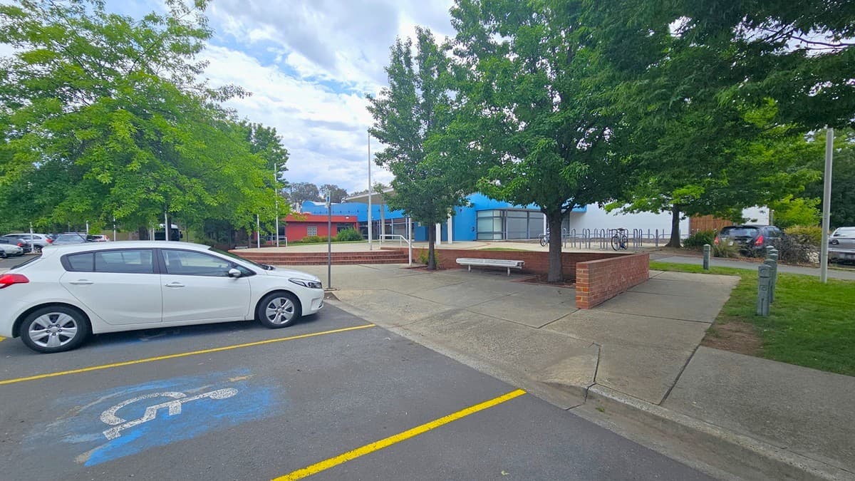 A white car is parked in a designated disabled parking space on the left, marked by blue paint and a wheelchair symbol. The ground is paved with yellow lines marking the parking area. In the middle, there are trees providing shade, and a brick bench is visible. In the background, a building with blue and red accents is partially obscured by trees. Additional cars are parked further back, and a bicycle rack is visible near the building.