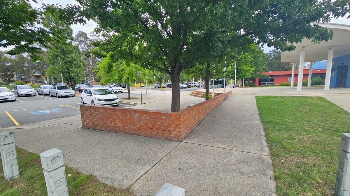 A paved area with a red brick wall runs through the centre, with a tree growing from it. On the left, there is a car park with several parked cars and yellow lines marking spaces. On the right, a grassy area leads to a building with a blue and red facade supported by white columns. The background features more trees and additional parked cars. The ground is a mix of concrete and grass.