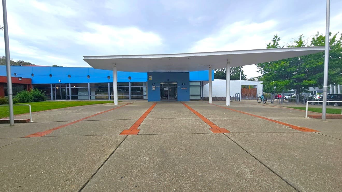 A building with a blue facade and a large white overhang supported by pillars. The entrance is centred, with glass doors and windows. The ground is paved with concrete, featuring red brick lines. On the left, there's a grassy area with shrubs, and on the right, trees and a bicycle rack. The background includes parked cars and more greenery.
