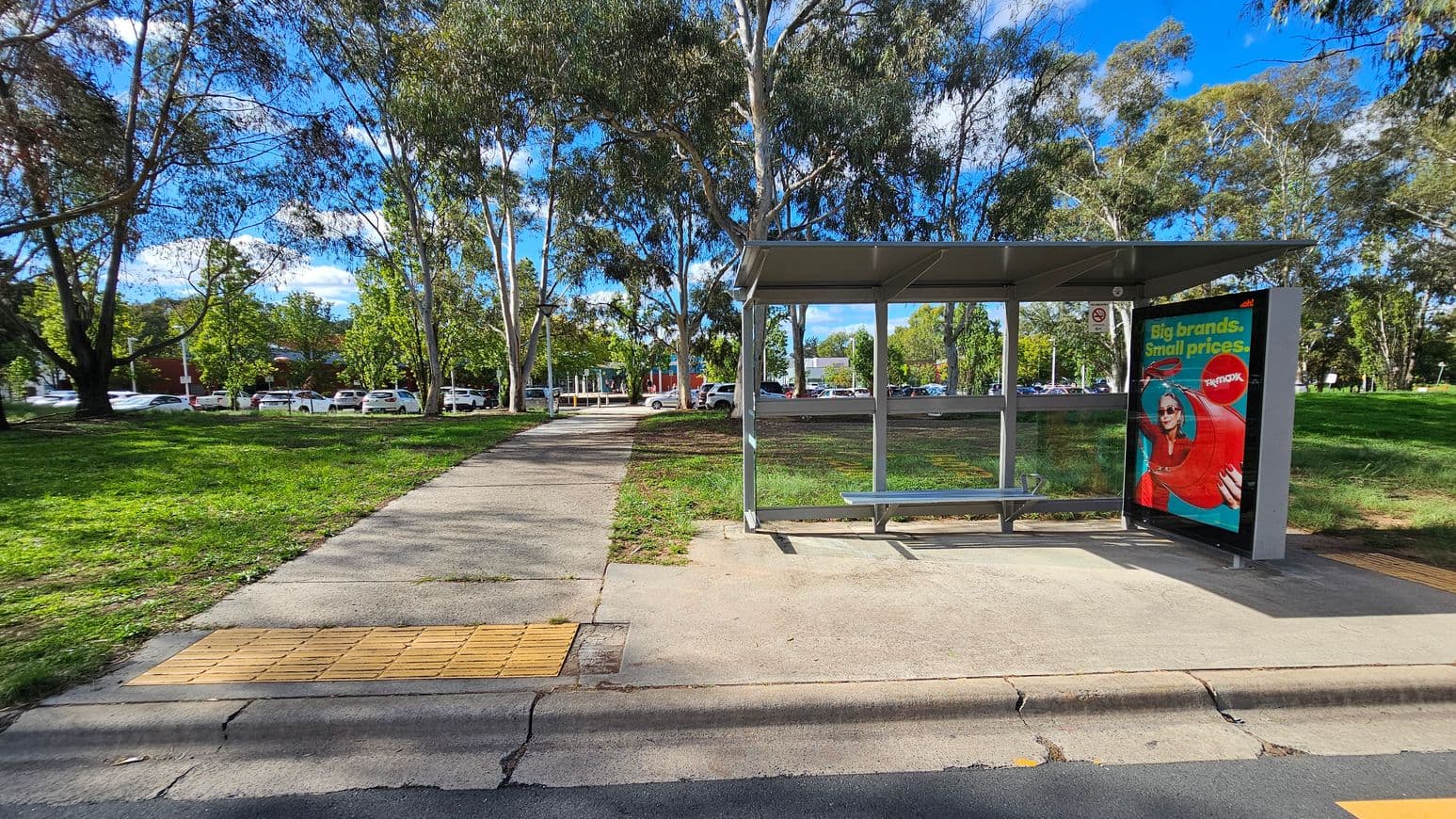 A bus stop with a metal shelter and a bench is on the right. An advertisement is displayed on the side of the shelter. A concrete path leads from the foreground to the background, flanked by grass and trees. In the background, there are parked cars and more trees. The sky is visible through the tree branches.