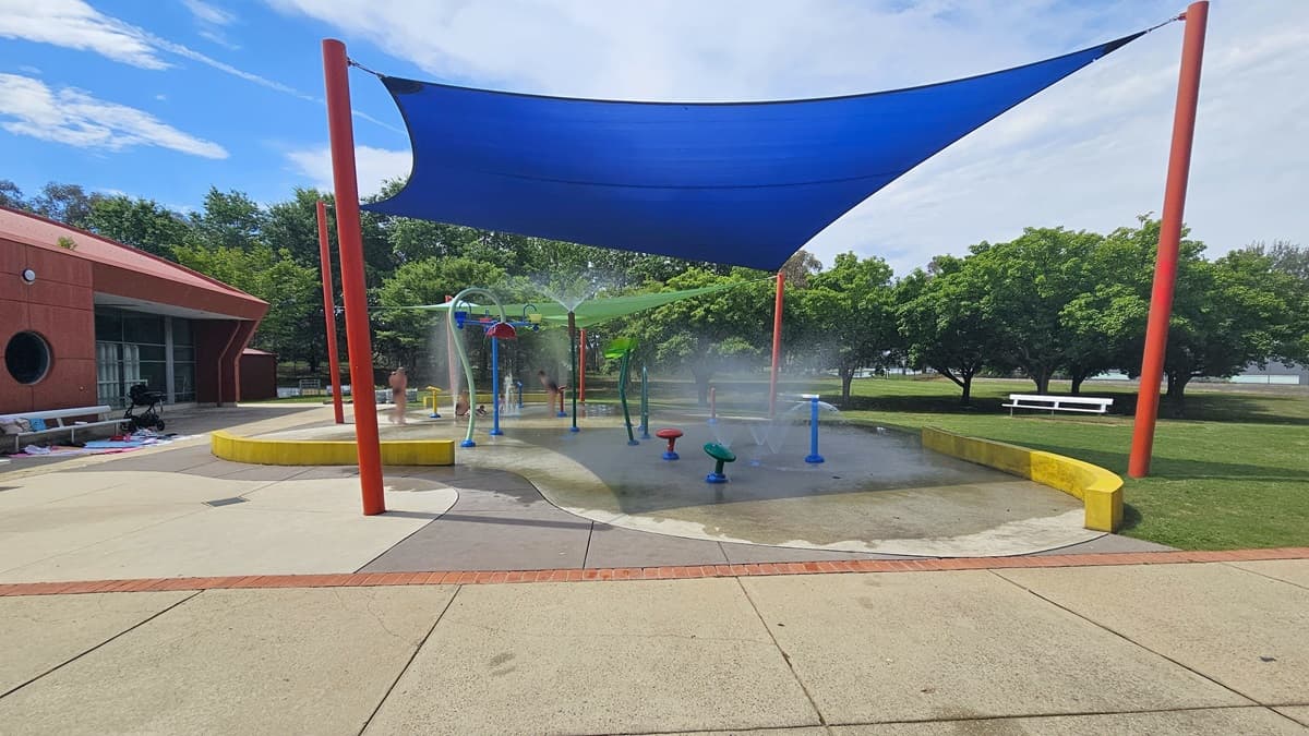 A splash pad with various water features is covered by a large blue shade sail supported by red poles. The ground is a mix of concrete and wet surfaces. On the left, a brick building with circular windows is visible. In the background, there are green trees and a white bench on a grassy area. The sky is partly cloudy.