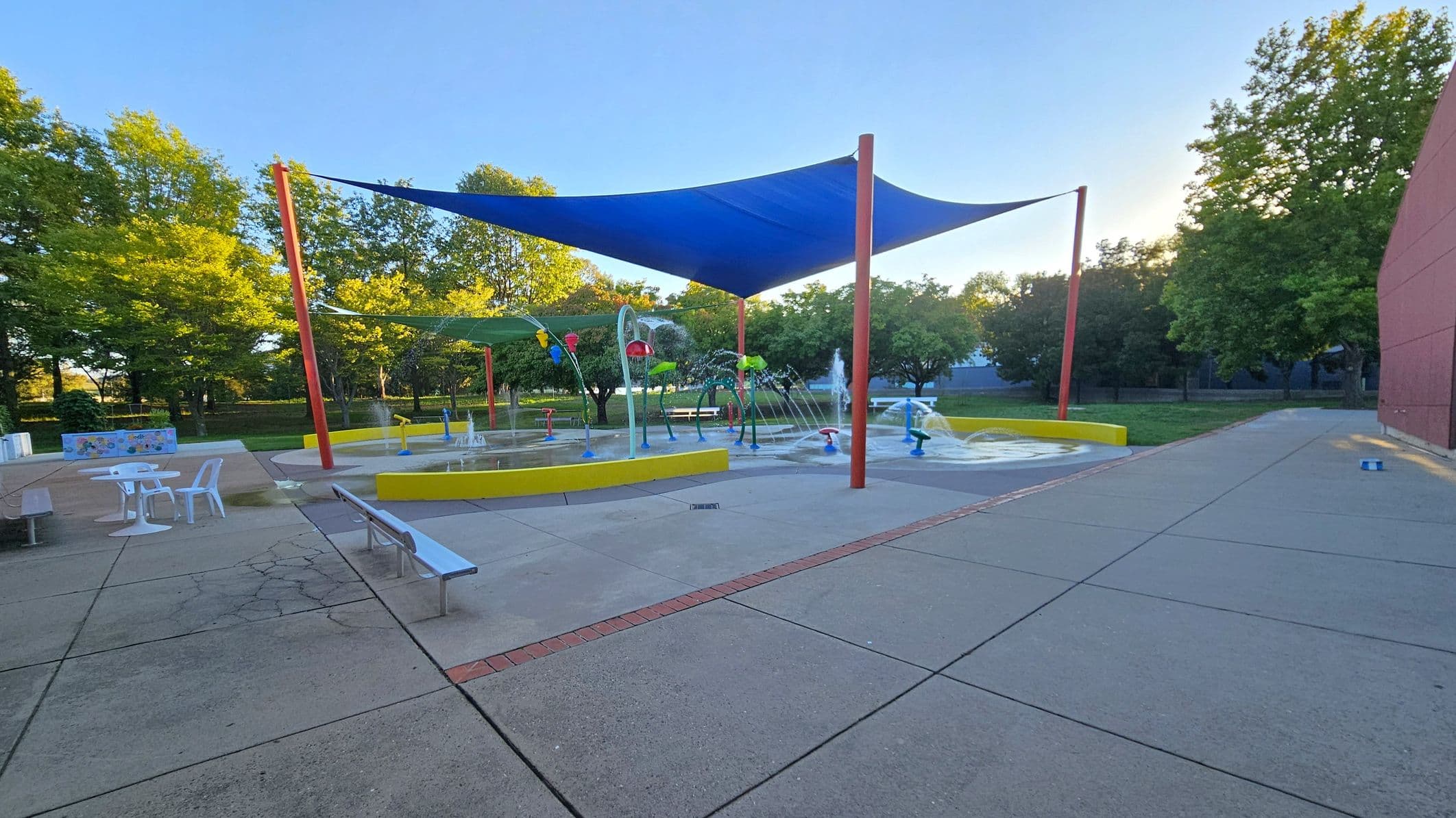 A splash pad with various water features is in the centre, surrounded by a yellow barrier. Blue and green shade sails are supported by red poles above. On the left, there are white tables and chairs on a paved area. The right side has a red brick wall and more pavement. Trees with green foliage are in the background, providing a natural setting. The ground is a mix of concrete and brick patterns.