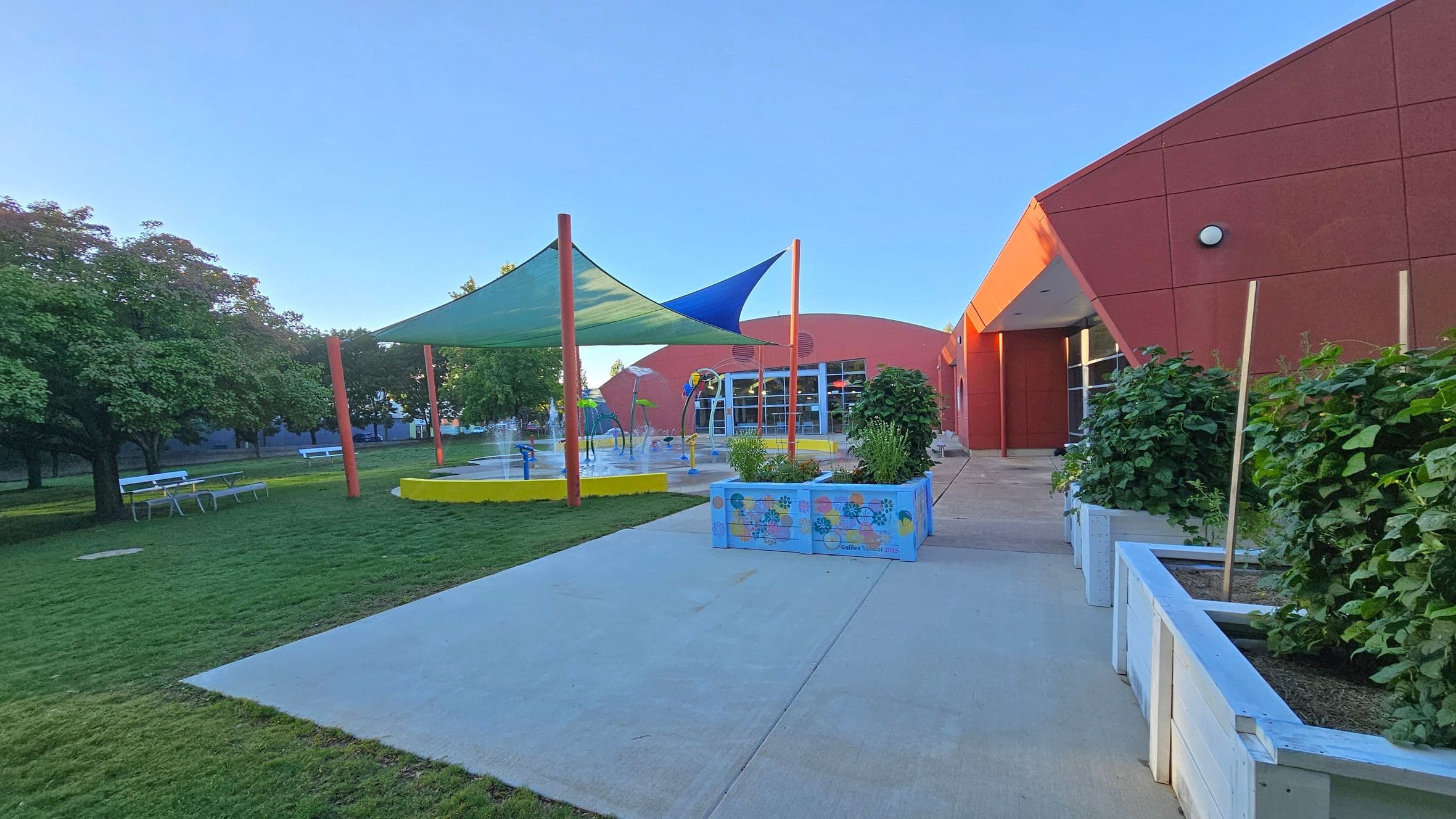 A playground with a splash pad is on the left, featuring a yellow circular base and colourful shade sails. Trees and picnic tables are in the grassy area. On the right, a red building with angular architecture and large windows is visible. In the foreground, there are planter boxes with greenery along a concrete path.