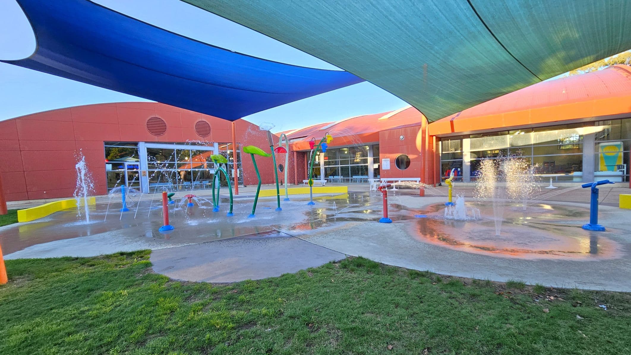 A splash pad with various water features, including fountains and colourful structures, is in the centre. The ground is wet, with some grass visible in the foreground. The background shows a red building with large windows and circular vents. Overhead, blue and green shade sails provide cover. The area is open and designed for recreational water play.