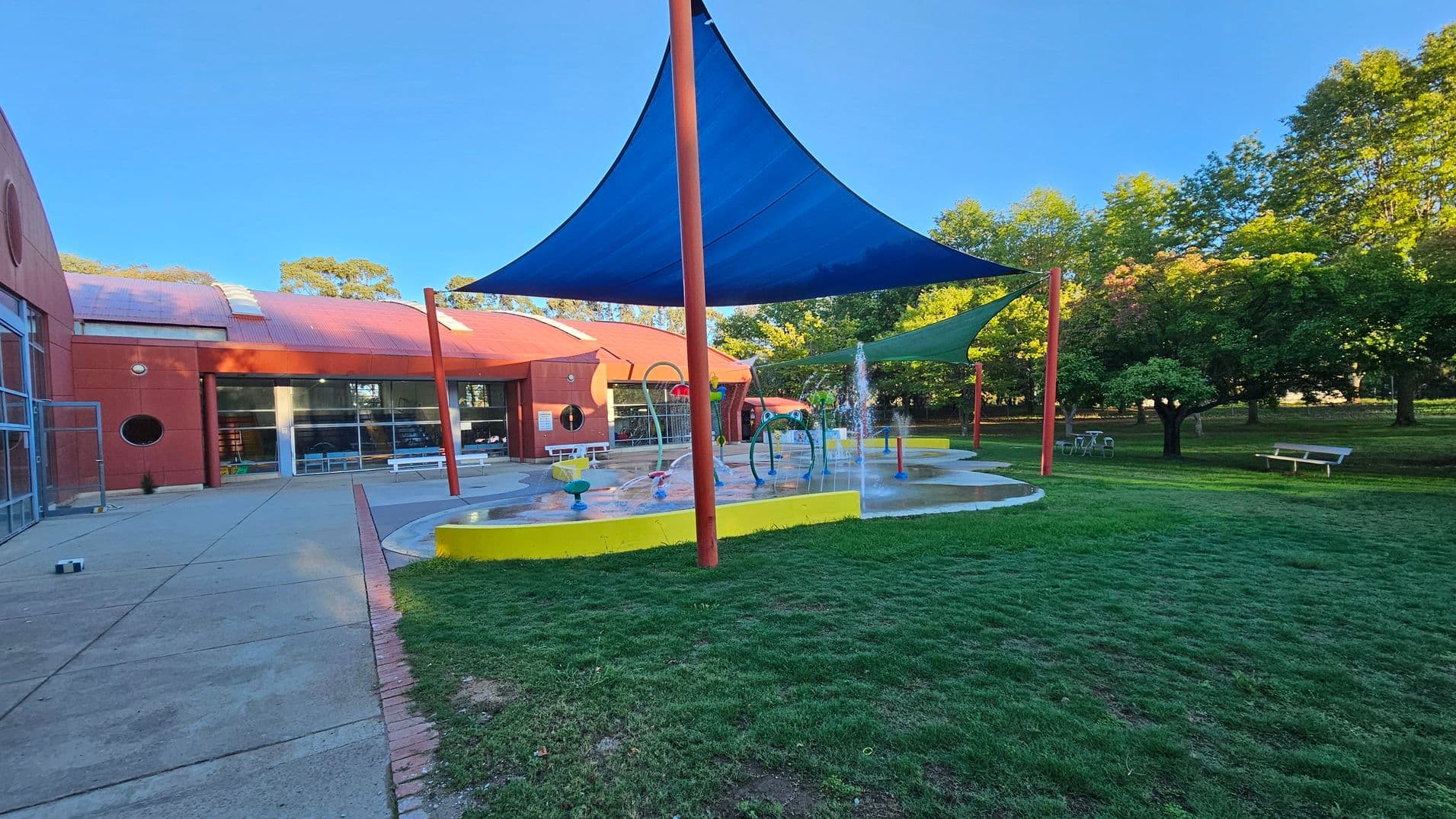 A playground with a splash pad is in the centre, featuring water fountains and colourful play structures. Blue and green shade sails are supported by red poles. On the left, a red brick building with large windows and a circular window is visible. The foreground has a grassy area, while the background includes trees and a bench. The ground is a mix of concrete and grass.