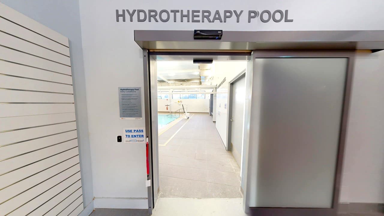 Entrance to a hydrotherapy pool area. The door is metallic with a sign above reading "HYDROTHERAPY POOL". To the left, there is a wall with horizontal lines and a sign instructing to use a pass to enter. Inside, a pool is visible with a pathway leading to it. The ceiling has exposed pipes and lighting fixtures.