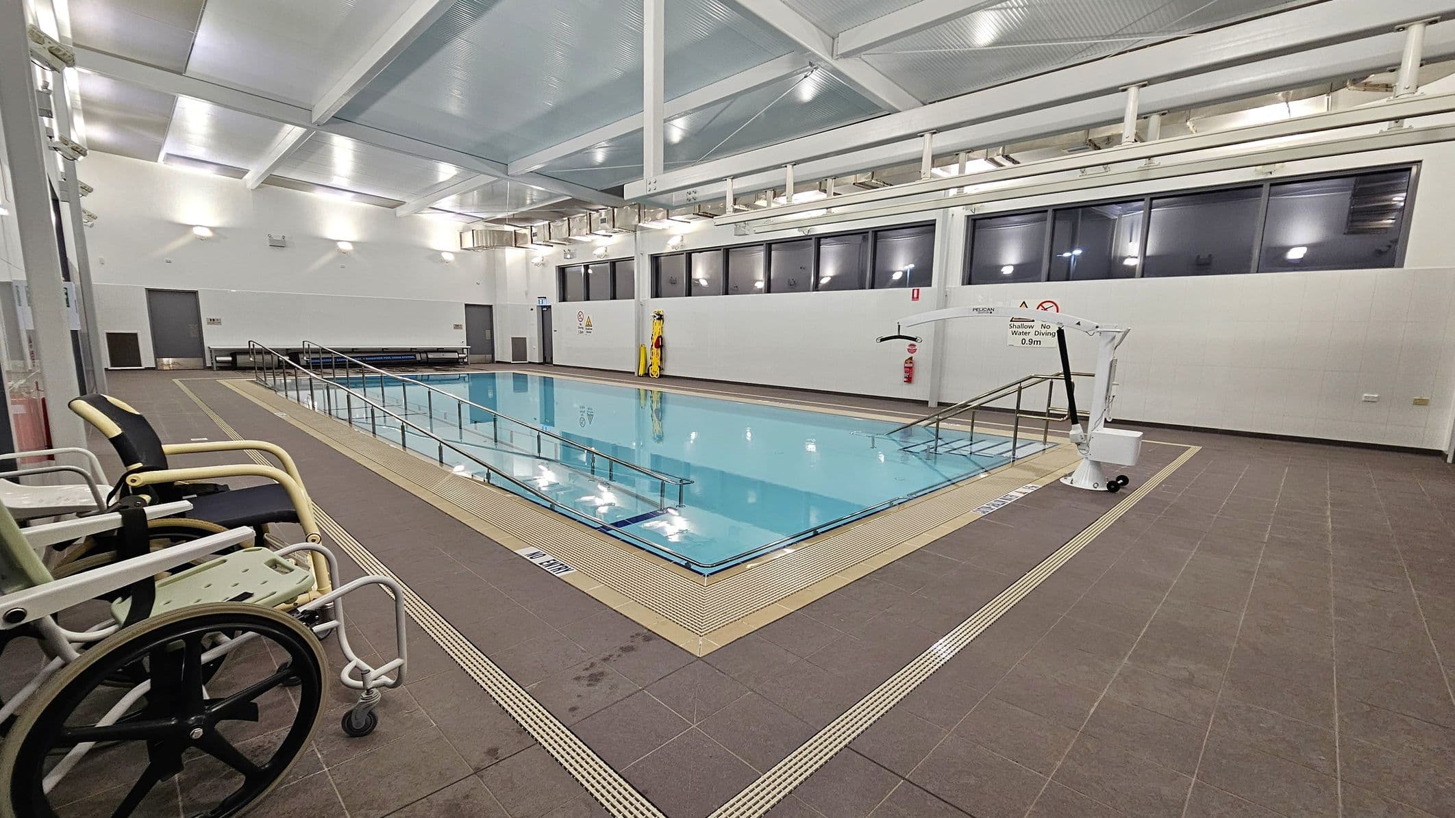 An indoor swimming pool with a ramp leading into the water, surrounded by a brown tiled floor. On the left, there are two wheelchairs positioned near the pool's edge. The right side features a mechanical pool lift. The back wall has large windows and a few doors, with a bench and safety equipment visible. The ceiling is high with exposed beams and bright lighting.