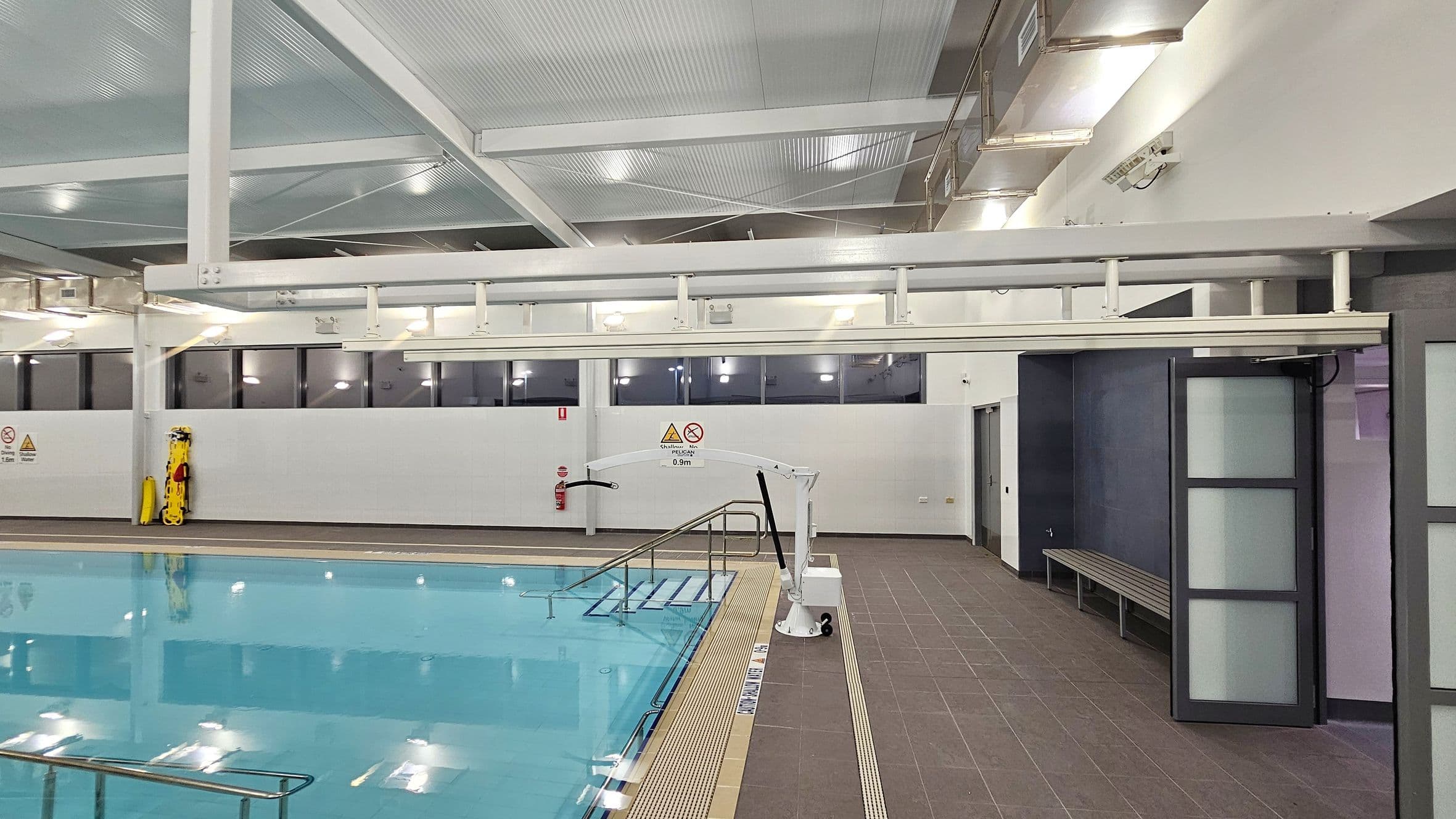 Indoor swimming pool area with a clear blue pool on the left. A metal handrail descends into the water. On the right, a brown tiled floor leads to a wall with a bench and a partially open door with frosted glass panels. Above, a white ceiling with exposed beams and lights. A pool lift is positioned near the pool edge. Windows line the upper wall, and a yellow safety board is mounted on the wall. Warning signs are visible near the pool.