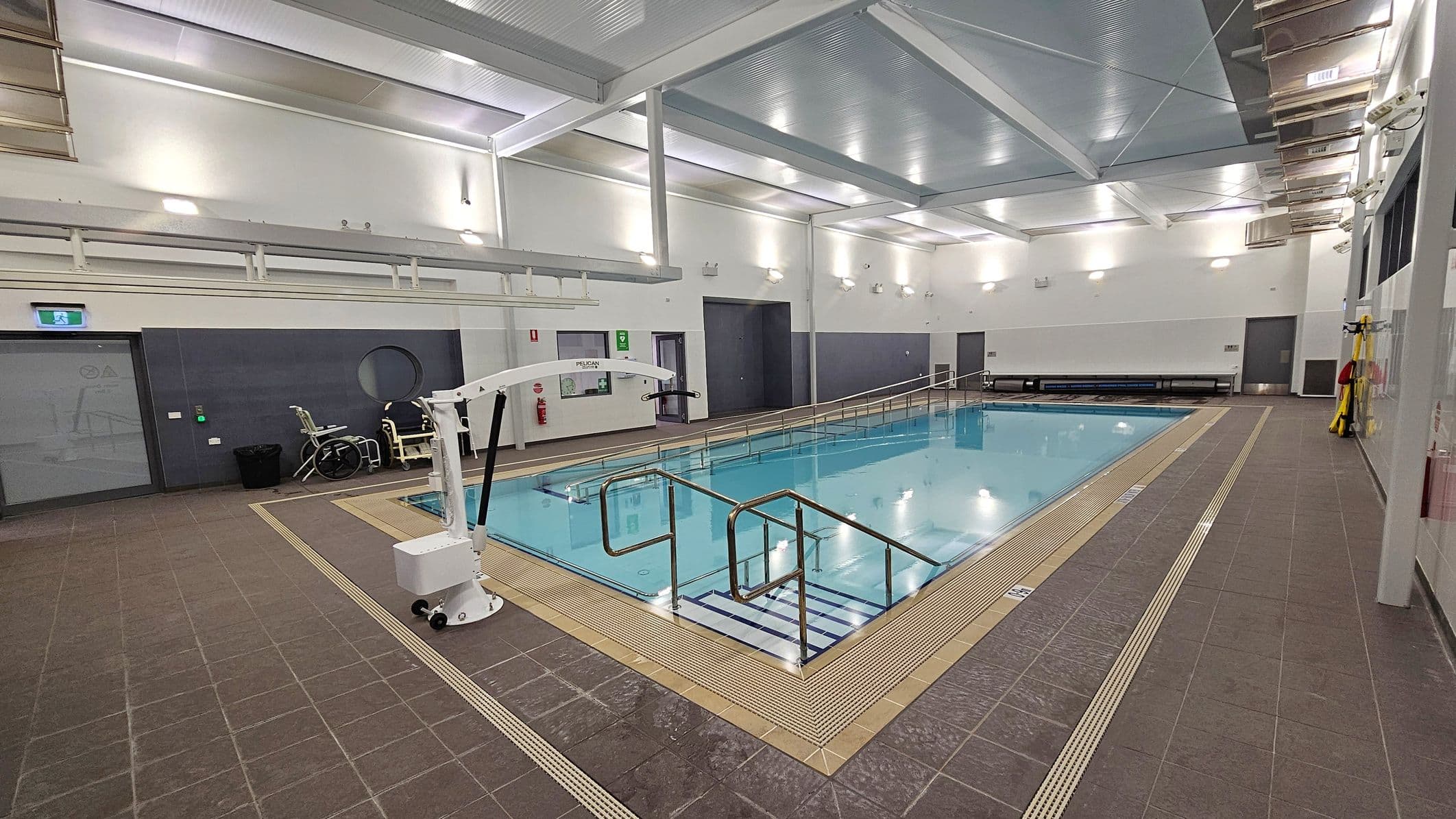 Indoor swimming pool with a tiled deck surrounding it. On the left, there's a pool lift and a wheelchair near a grey door with a circular window. The pool has metal handrails and a ramp for accessibility. The walls are white with several lights, and there are benches along the far wall. The ceiling is high with exposed beams and bright lighting. The floor is brown tile with drainage grates around the pool's edge.
