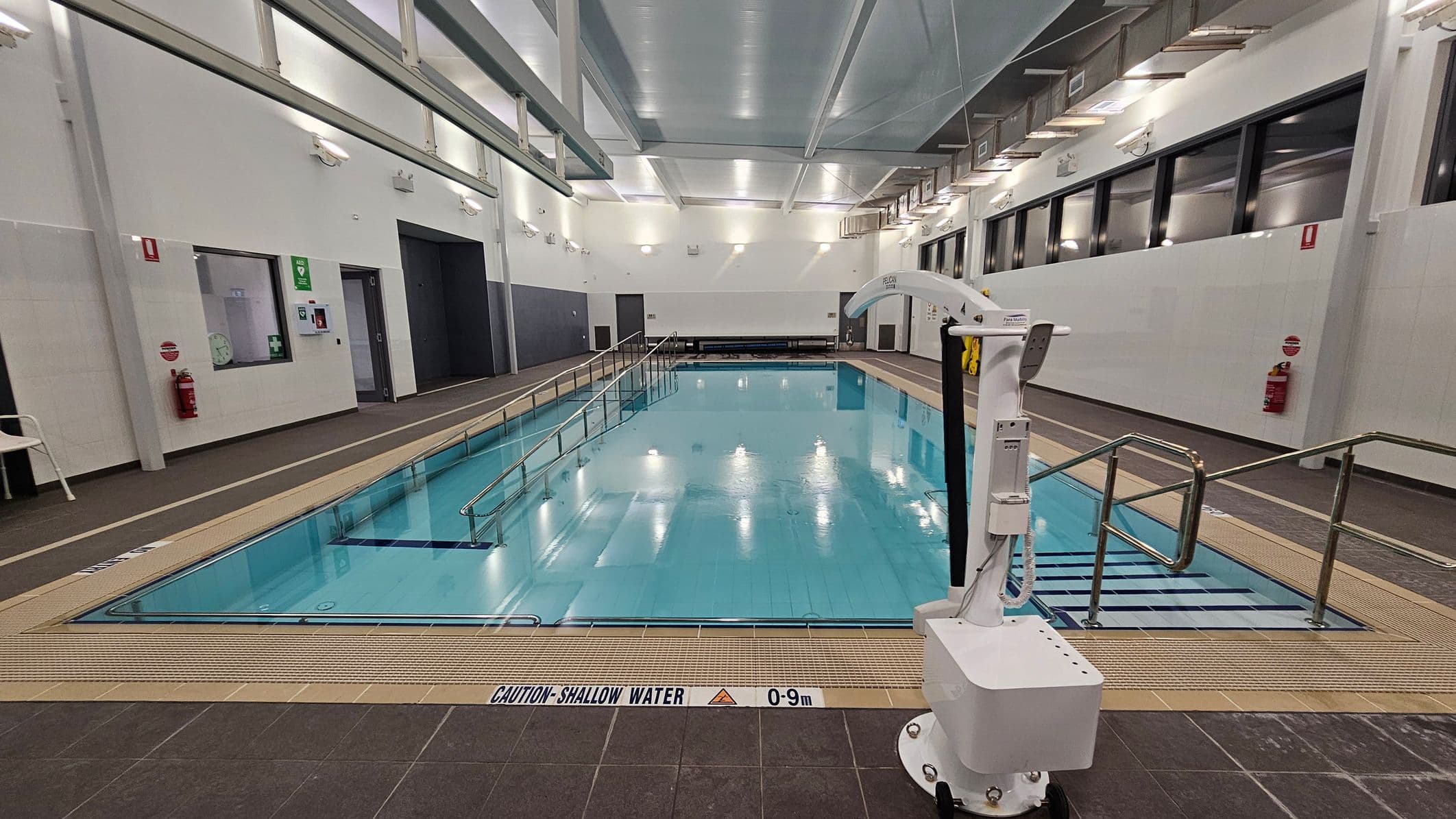 An indoor swimming pool with clear blue water is in the centre. On the right, a white pool lift is positioned near metal handrails leading into the pool. The left side features a wall with emergency equipment, including a fire extinguisher and a first aid kit. The ceiling is high with exposed beams and lights. The floor surrounding the pool is tiled in dark brown, with a caution sign indicating shallow water. Large windows line the right wall.