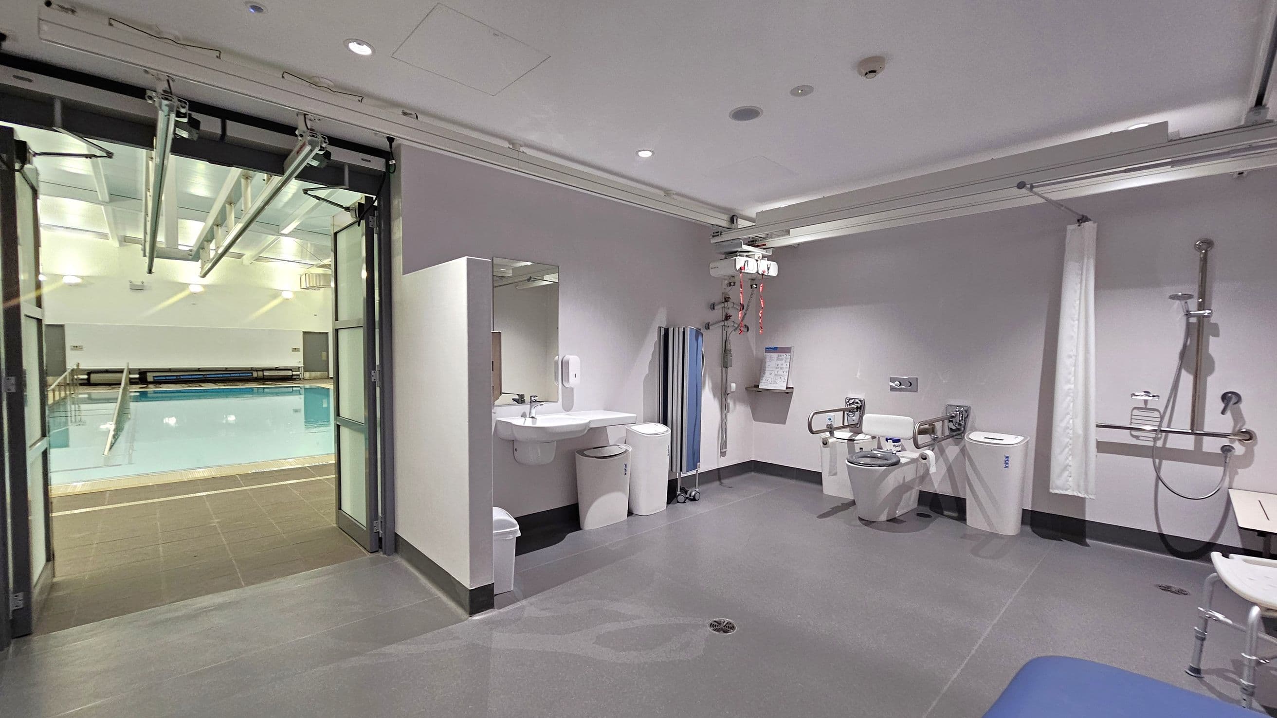 On the left, there is a swimming pool visible through large glass doors. The pool area is well-lit with overhead lights. In the middle, a bathroom area features a sink with a mirror, soap dispenser, and bins underneath. On the right, there is a toilet with support rails, a shower with a handheld nozzle, and a shower chair. The ceiling has recessed lighting and a track system for a hoist. The floor is smooth and grey.