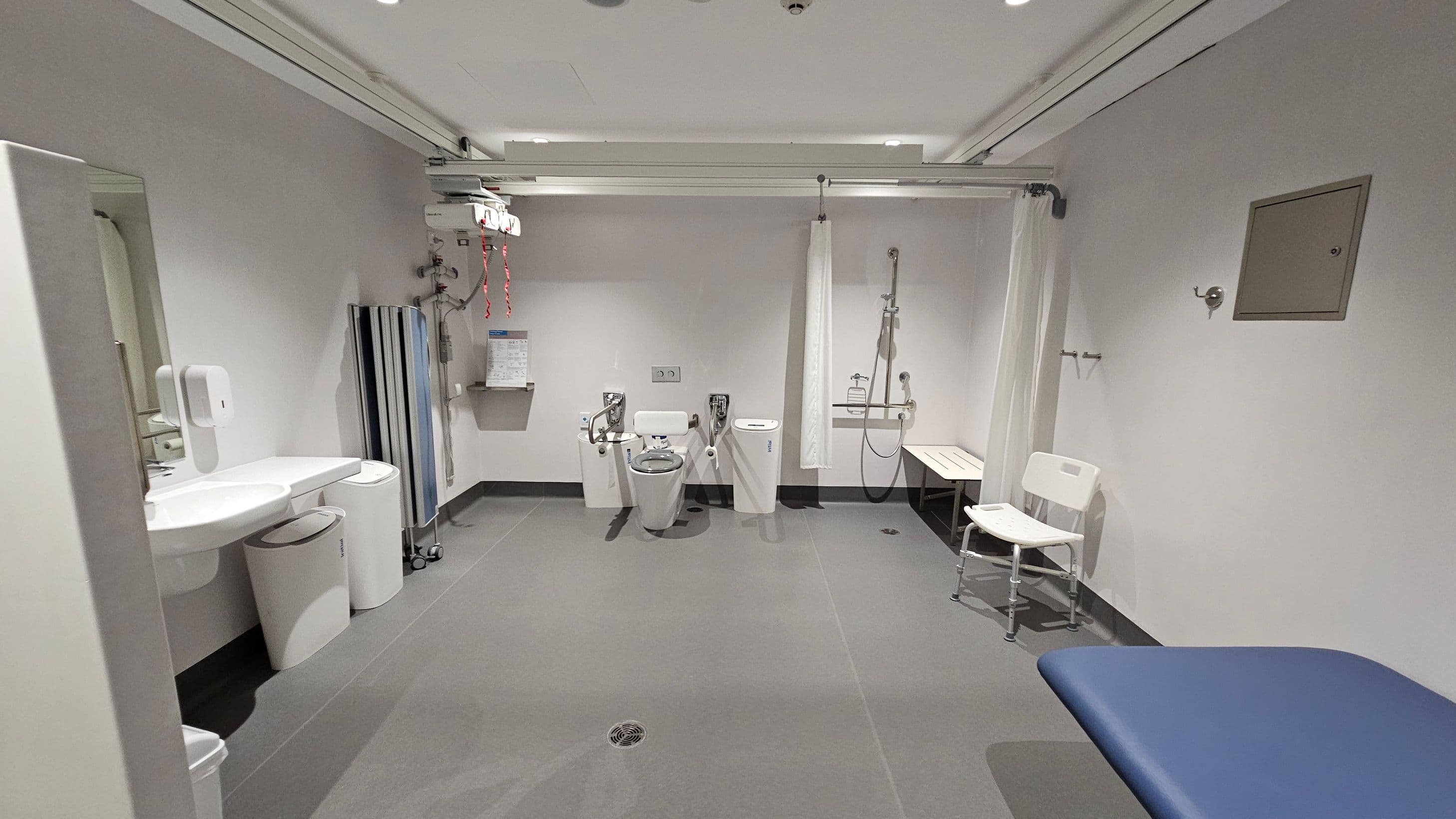 A spacious, accessible bathroom with grey tiled flooring and a white ceiling. On the left, there is a sink with a soap dispenser and bins underneath. In the centre, a toilet with support rails is visible, along with a wall-mounted hoist system. To the right, a shower area with a curtain, a bench, and a white chair is present. A blue padded bench is in the foreground on the right. The walls are plain with a small notice board on the right.