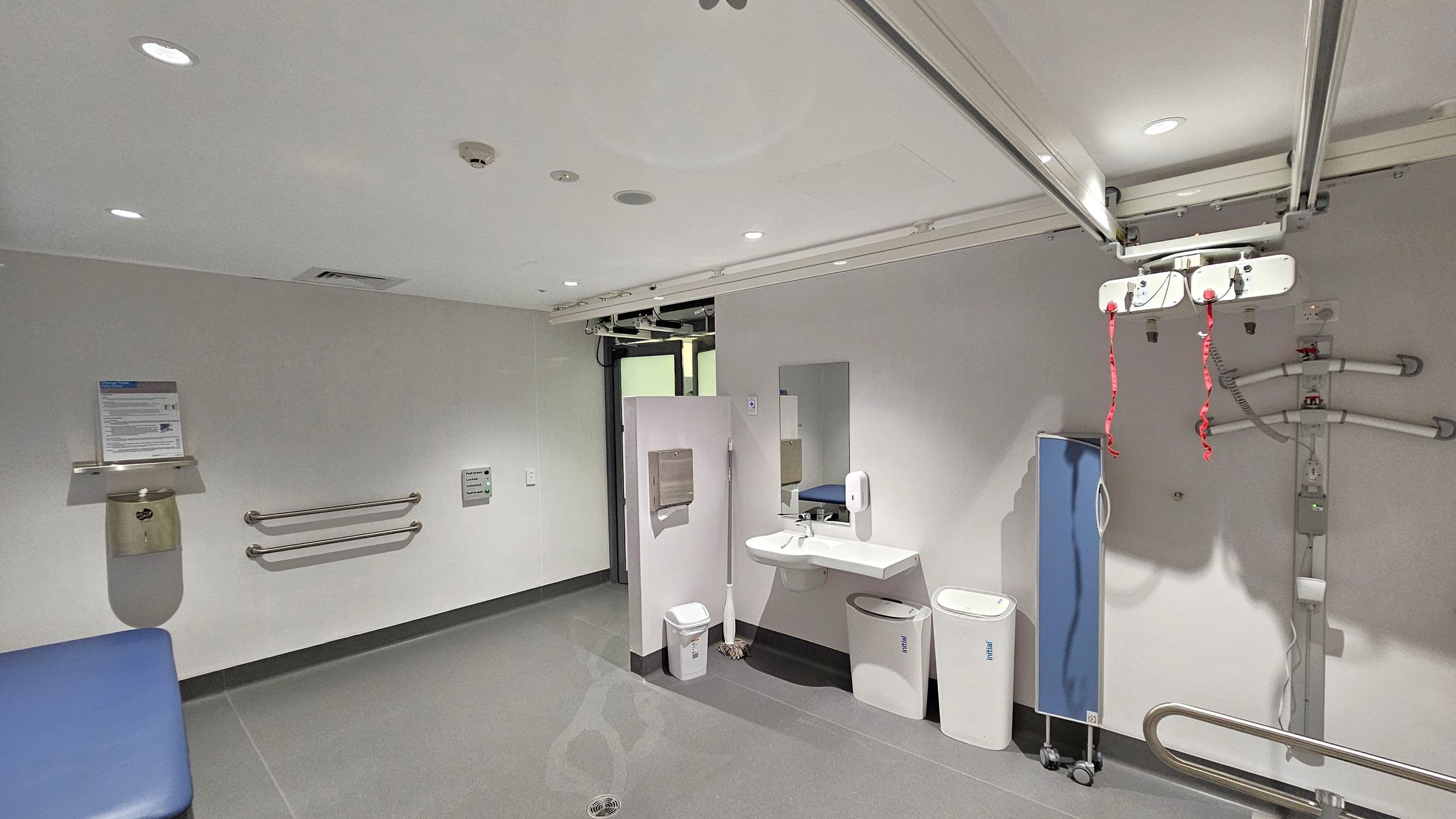 A hospital room with a grey floor and white ceiling. On the left, there are two horizontal grab bars and a wall-mounted bin with a sign above it. In the centre, a sink with a mirror, soap dispenser, and two bins below. A mop is leaning against the wall. On the right, a blue privacy screen on wheels and a ceiling-mounted hoist with red straps. The room has recessed lighting and a door in the background.