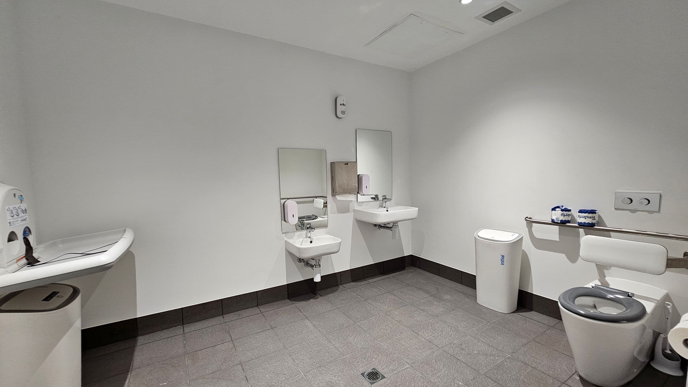 A public restroom with grey tiled flooring and white walls. On the left, a baby changing station is mounted above a bin. In the centre, two wall-mounted sinks with mirrors and soap dispensers are visible. On the right, a toilet with a support bar and two rolls of toilet paper on top. A sanitary bin is placed beside the toilet. The ceiling is plain with recessed lighting.