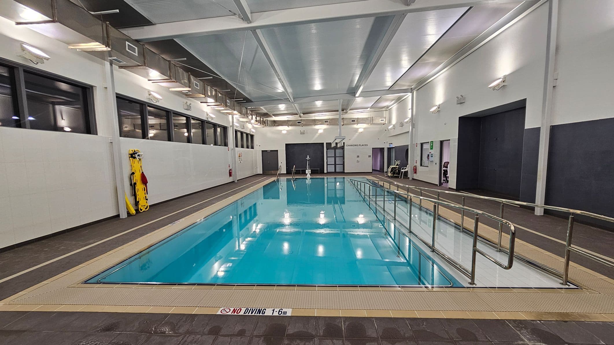 An indoor swimming pool with clear blue water is in the centre. On the left, there are large windows and a yellow rescue board against the wall. On the right, there is a ramp with metal railings leading into the pool. The walls are white with several lights, and there are doors labelled "Changing Places" in the background. The ceiling is high with exposed beams and ventilation ducts. The floor around the pool is tiled in a dark colour.