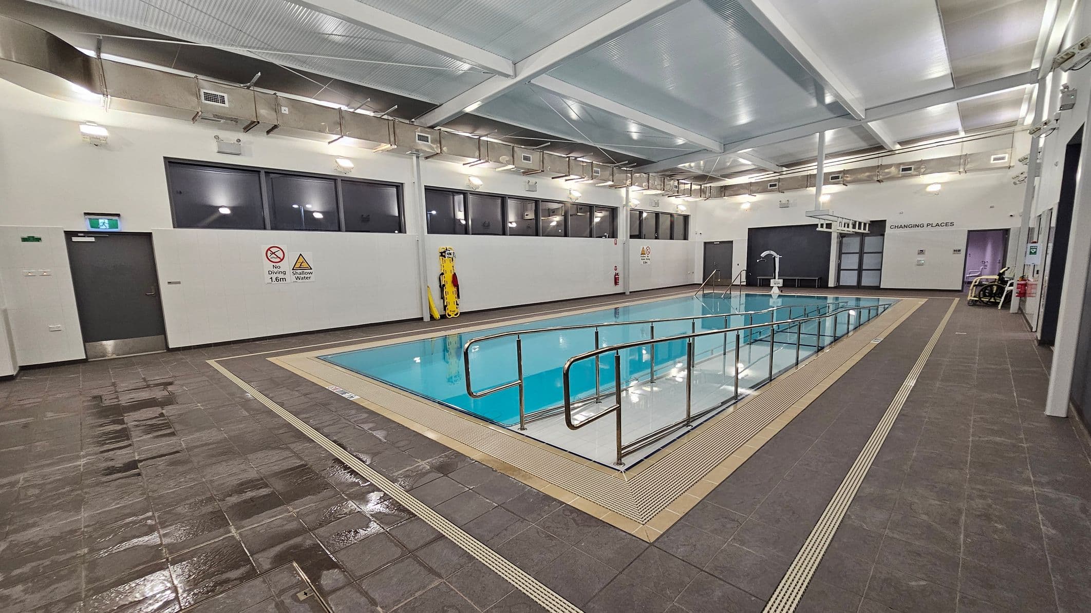 An indoor swimming pool with a metal handrail along its edge. The pool is surrounded by wet, brown tiles. On the left wall, there are signs indicating "No Diving" and "Shallow Water," along with a door and a mounted rescue board. The right side features a door labelled "Changing Places" and a wheelchair. The ceiling is high with exposed beams and lighting fixtures. Large windows line the upper part of the walls.