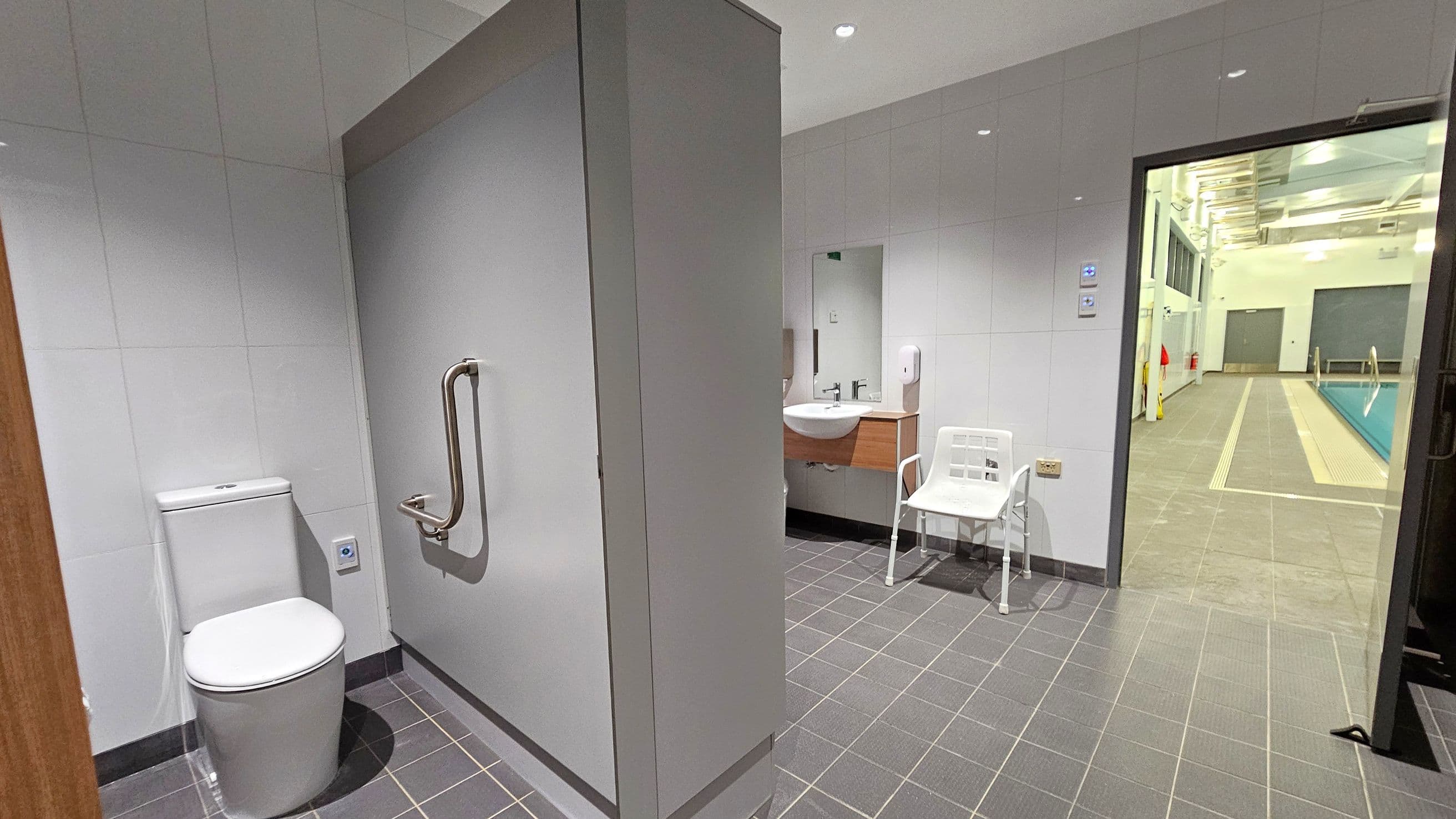 A bathroom with grey tiled flooring and white tiled walls. On the left, there is a toilet with a metal grab bar. In the centre, a sink with a mirror above it and a white chair nearby. On the right, an open door leads to a swimming pool area with a tiled walkway and a pool visible. The ceiling has recessed lighting.