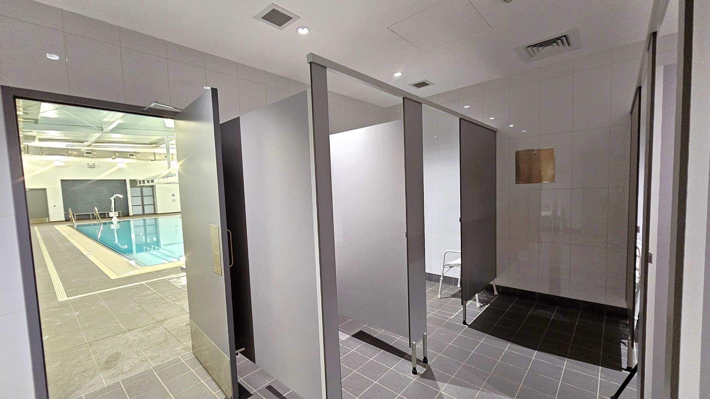 On the left, there is a view of an indoor swimming pool with a tiled deck and metal handrails. The pool area is well-lit with overhead lights and has a high ceiling. On the right, there are changing cubicles with grey doors and tiled flooring. A small white chair is visible inside one of the cubicles. The ceiling is white with recessed lighting and ventilation panels.