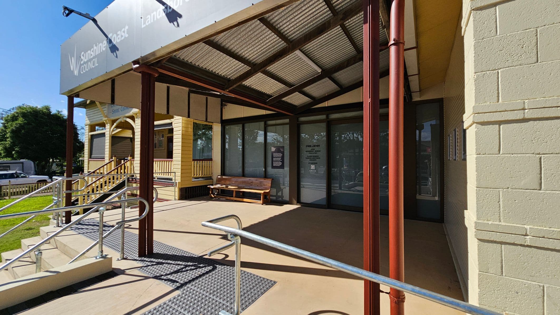 A building entrance with a covered porch supported by brown metal poles. The porch has a sloped roof with a metal frame and corrugated panels. A wooden bench is placed against the glass doors on the right. A ramp with metal railings leads up to the entrance. On the left, a yellow wooden structure with a small staircase and railing is visible. The ground is concrete, and there is a grassy area with a tree in the background.