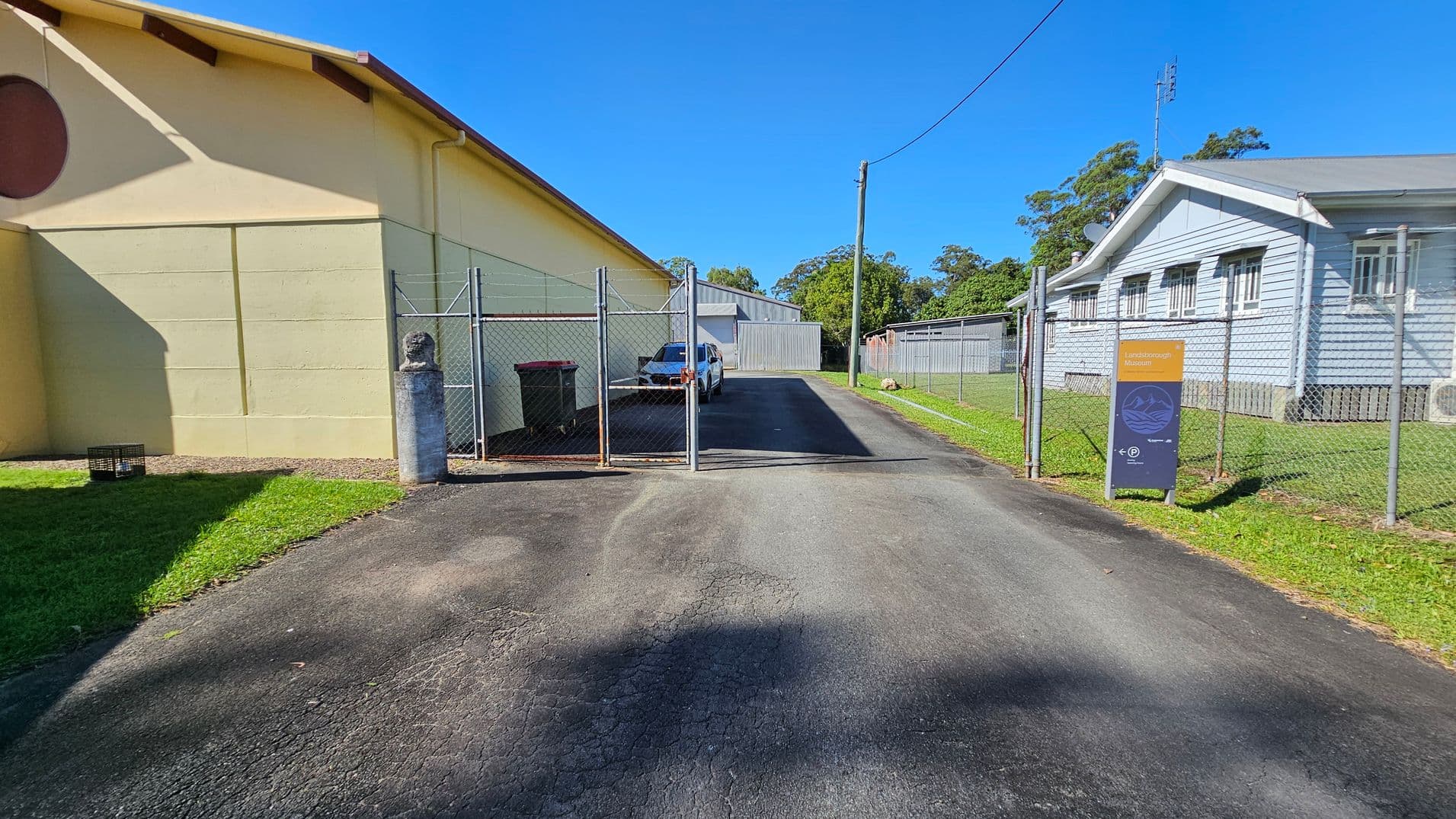 A narrow asphalt driveway leads to a fenced area with a gate. On the left, a large beige building casts a shadow on the grass. A white vehicle is parked inside the fenced area. On the right, a light blue building with multiple windows runs parallel to the driveway. A sign with text is positioned near the fence on the right side. Trees and a utility pole are visible in the background.