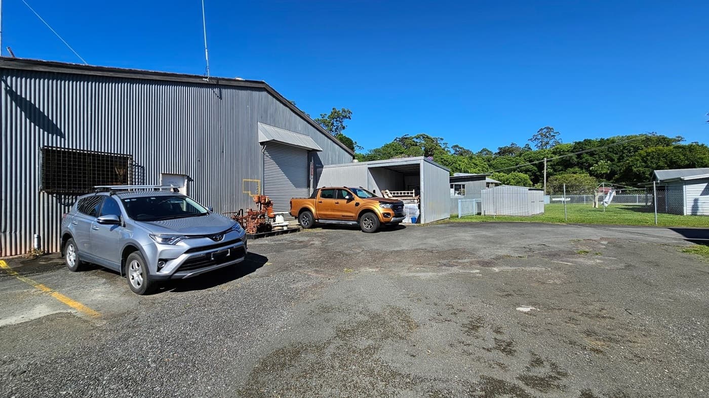 A corrugated metal building with a grey exterior is on the left, with a silver SUV parked in front. Next to it, a golden pickup truck is parked near a small structure with a roller door. The ground is a mix of gravel and patches of grass. In the background, there are more metal structures and a chain-link fence enclosing a grassy area with trees beyond.