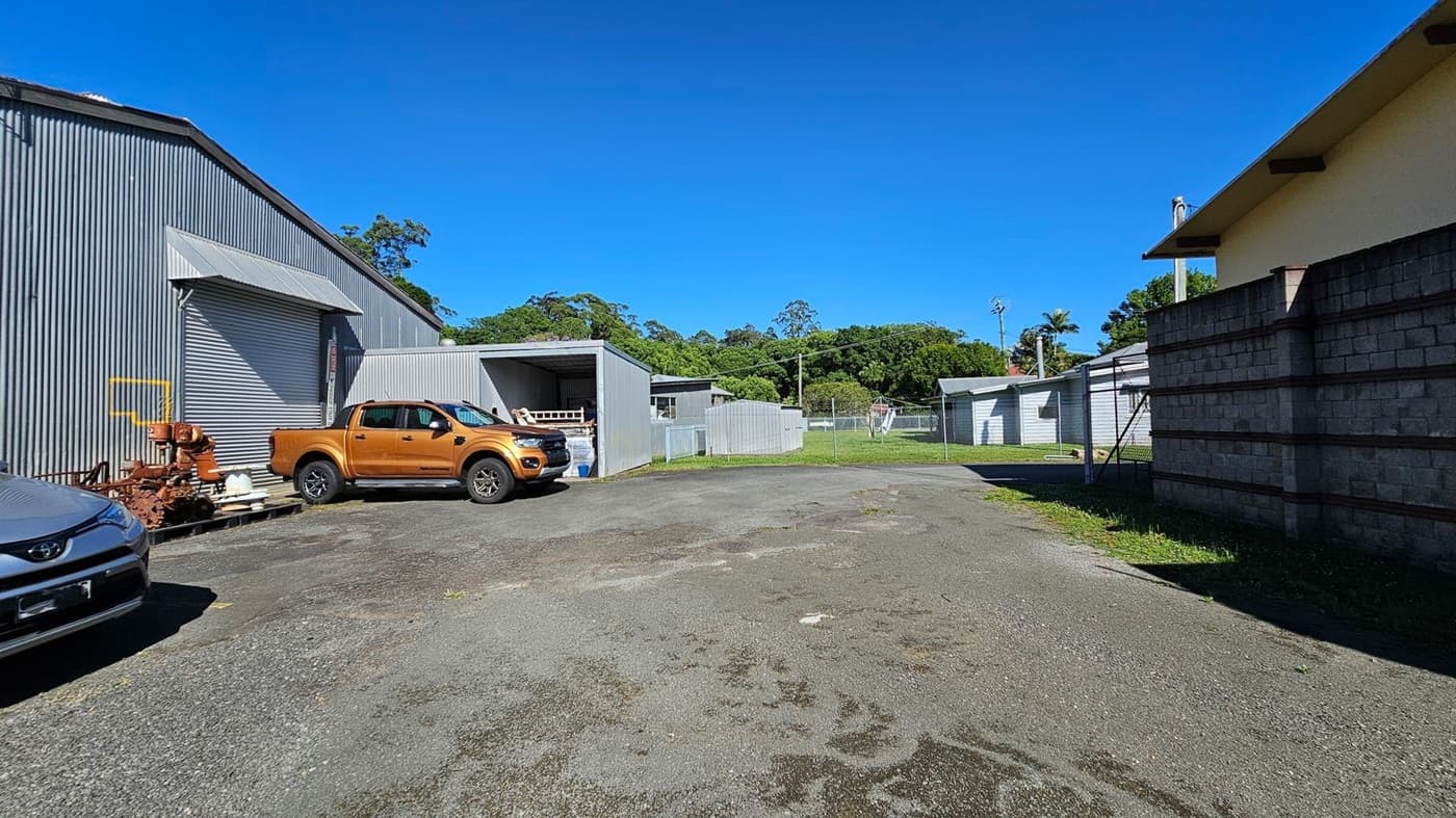 A paved area with patches of grass and moss. On the left, there's a large corrugated metal building with a closed roller door, next to which is a rust-coloured pickup truck. A grey car is partially visible in the foreground. In the centre, a small open shed and a fenced grassy area are visible. On the right, a brick wall and a smaller building with a sloped roof. Trees and greenery are in the background.