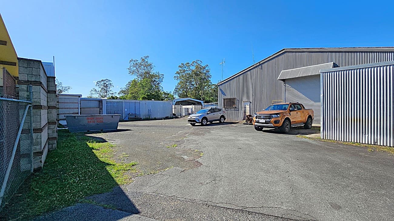 A paved industrial yard with a corrugated metal building on the right. Two vehicles, a silver SUV and an orange pickup truck, are parked in front of the building. On the left, a chain-link fence and a brick wall are visible. In the centre, a large metal skip bin is placed on the ground. In the background, there are more metal structures and some trees. The sky is clear and blue.