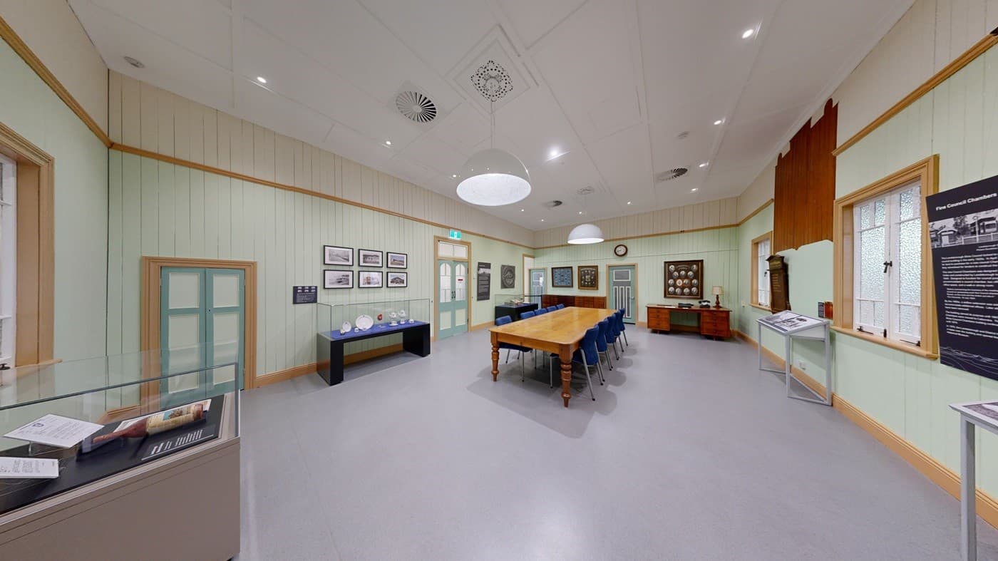 A spacious indoor area with a tiled floor and a wooden deck on the right, leading up to a building with cream-coloured weatherboard walls and large windows. The ceiling has exposed beams and metal trusses. On the left, there is a large frosted glass sliding door. Inside the building, tables and chairs are visible, and a sign marked "MACHINERY" points to the right. A small green pushchair is on the deck.