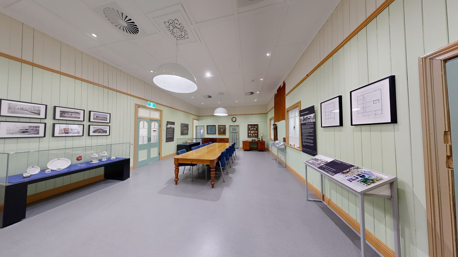 A room with a large wooden table surrounded by blue chairs on the left. The walls are light green with framed pictures and a large clock on the right. A door with a decorative design is also on the right. In the background, there are display cases with various items and a set of double doors. The ceiling has white panels with circular light fixtures and vents. The floor is light grey.